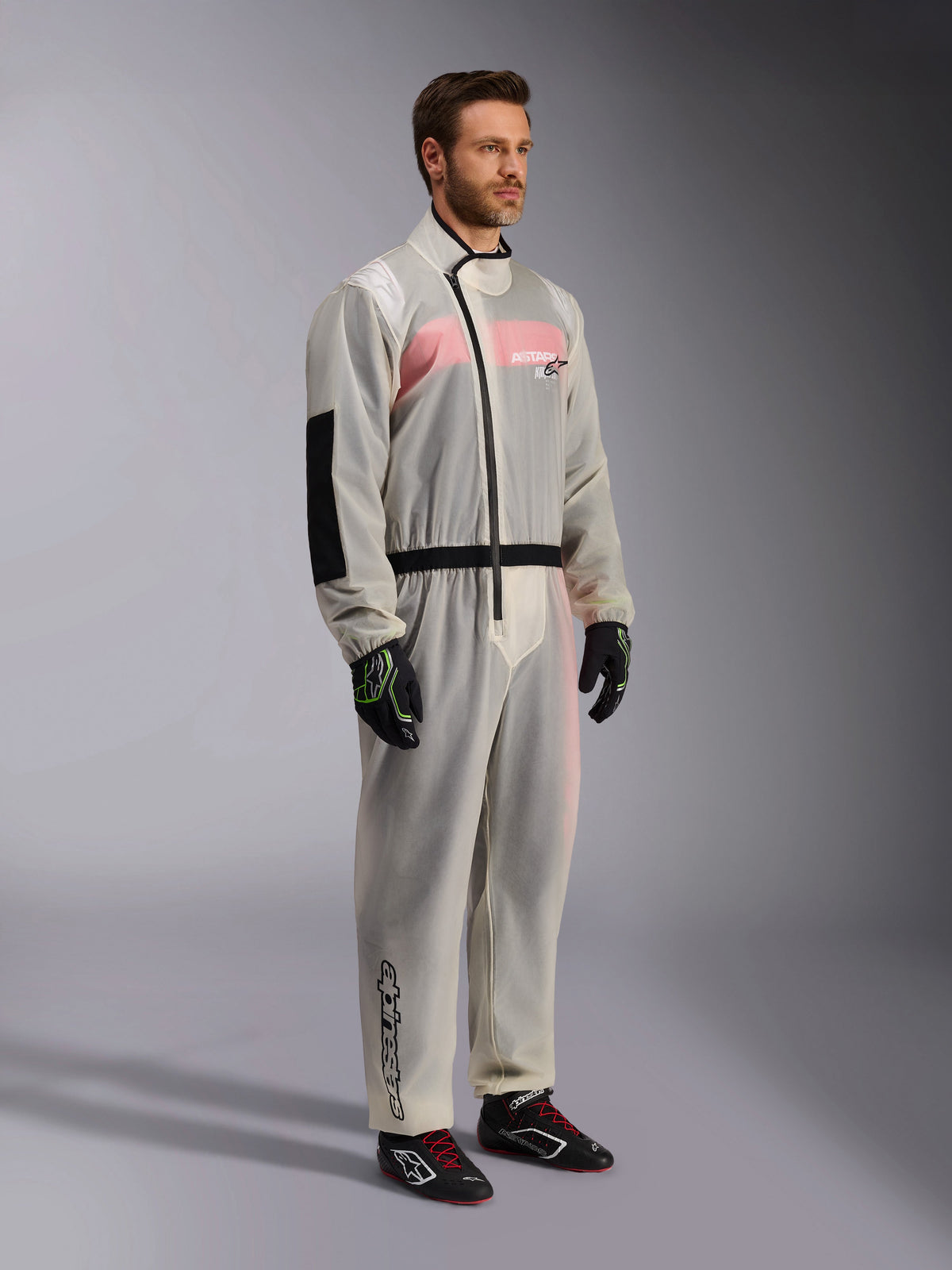 Alpinestars Kart Rain Suit, macacão de chuva para karting transparente e impermeável, material semitransparente que mostra um fato de corrida cor-de-rosa e branco por baixo, com fecho assimétrico preto, reforço no cotovelo e faixa elástica preta na cintura, usado com luvas de karting e sapatos de competição pretos