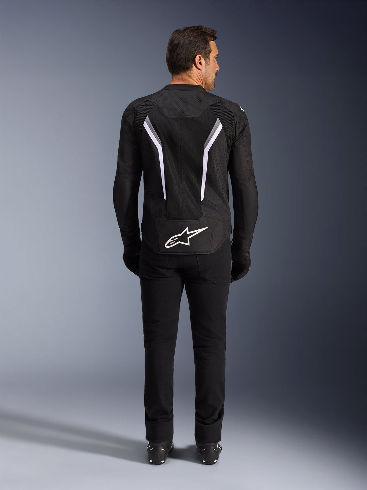 Casaco Alpinestars AST V3 Air, vista traseira, preto e cinzento escuro com detalhes brancos, com logótipo Astar grande a branco na parte inferior, riscas refletoras nas costas e construção em malha têxtil, usado com calças e botas pretas para um look de condução desportiva.