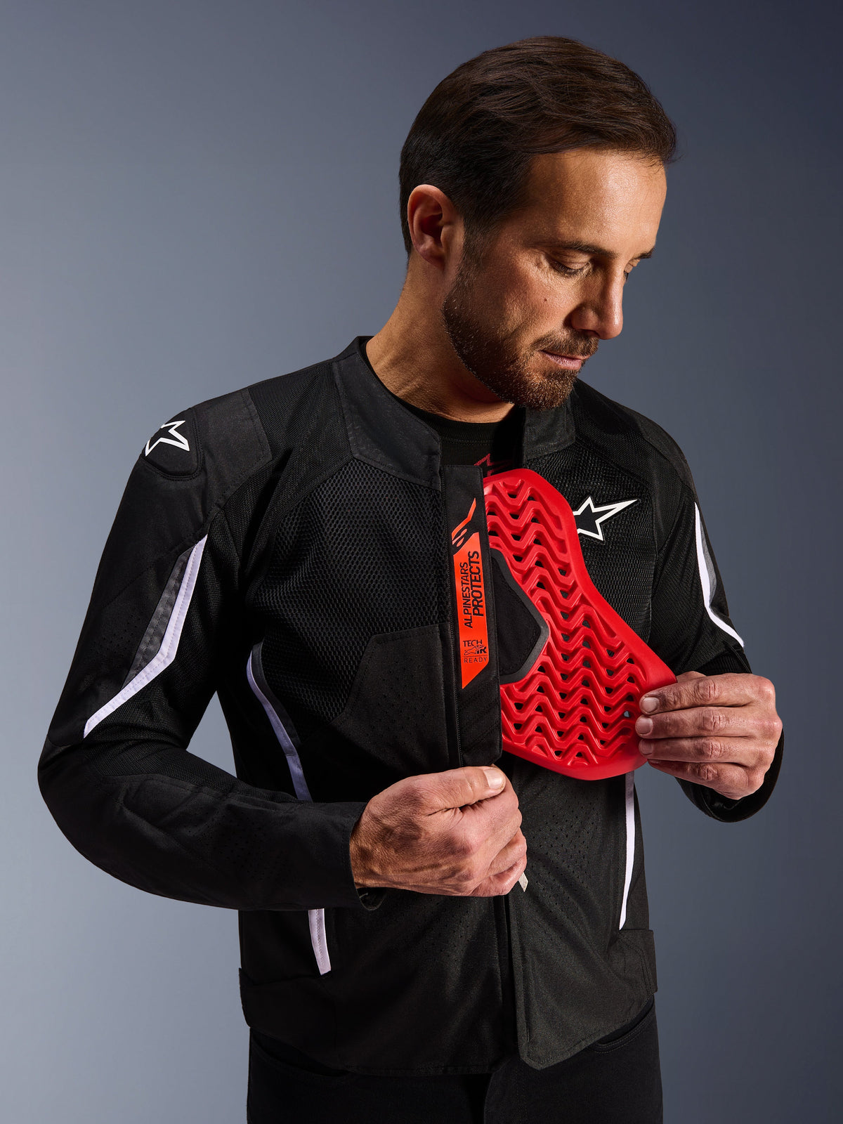 Alpinestars AST V3 Air Jacket em preto, homem a revelar um protetor de peito vermelho vivo com padrão em favo de mel, destacando a compatibilidade Tech-Air Ready e a armadura interna integrada para segurança em motociclismo