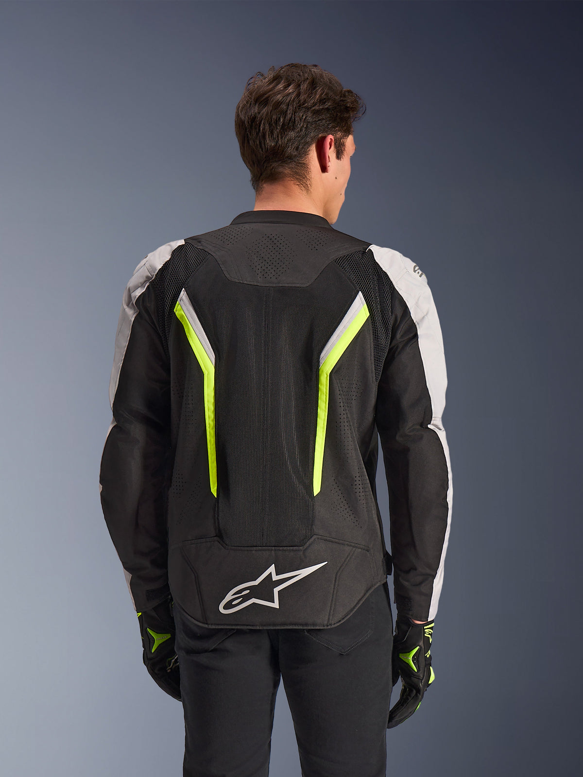 Alpinestars AST V3 Air Jacket, Black Silver Yellow Fluo, rear view on model, textile motorcycle jacket featuring dual tech mesh ventilation panels, vertical neon yellow stripes, silver sleeves, and a white Alpinestars logo on the lower back.