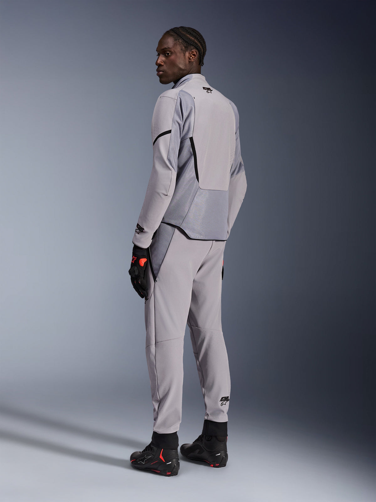 Casaco Alpinestars Aeroshell Airflow em Mid Gray Anthracite, usado com calças a condizer e luvas de corrida pretas, vista traseira que mostra os painéis perfurados a laser e o corte desportivo atlético, vestuário profissional para motociclismo
