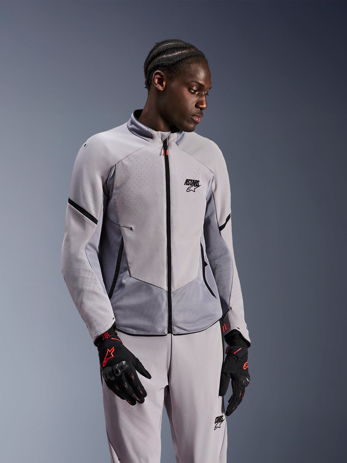 Casaco Alpinestars Aeroshell Airflow, Cinzento Médio Antracite, usado por um modelo com calças têxteis a condizer e luvas pretas com detalhes vermelhos, com zonas perfuradas a laser e painéis de malha para ventilação, design elegante de corte desportivo para condução de motociclos.