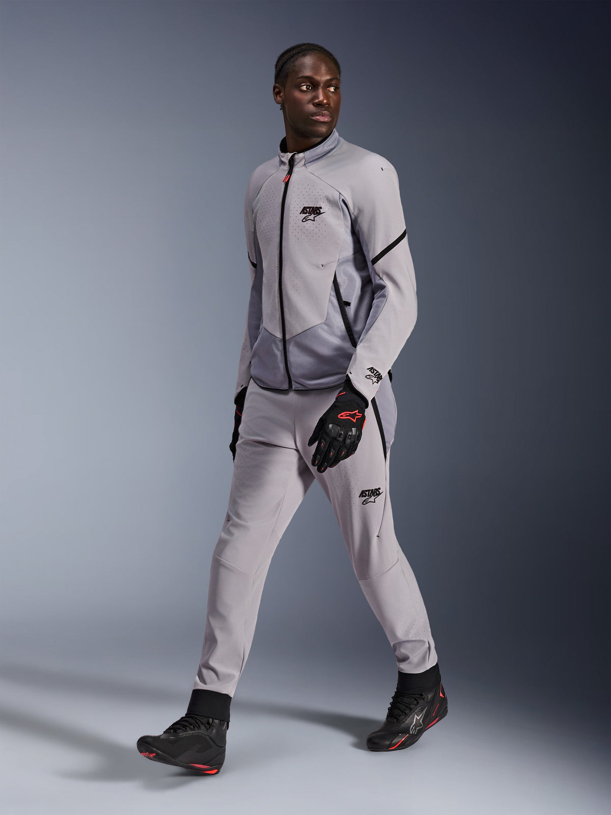 Casaco Alpinestars Aeroshell Airflow em cinzento Mid Gray Anthracite, usado por um modelo masculino com calças de condução cinzentas a condizer e luvas pretas, com ventilação no peito perfurada a laser e um design elegante de corte desportivo.