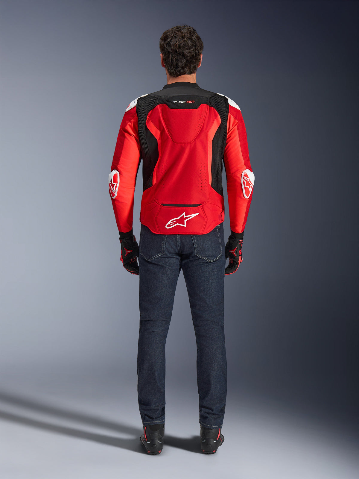 Casaco Alpinestars T-GP Air, vista traseira, vermelho vivo e preto, com logótipo branco na parte inferior das costas e marca 'T-GP AIR', usado com calças de ganga azul escuro e luvas de condução pretas e vermelhas, equipamento de motociclismo de performance desportiva