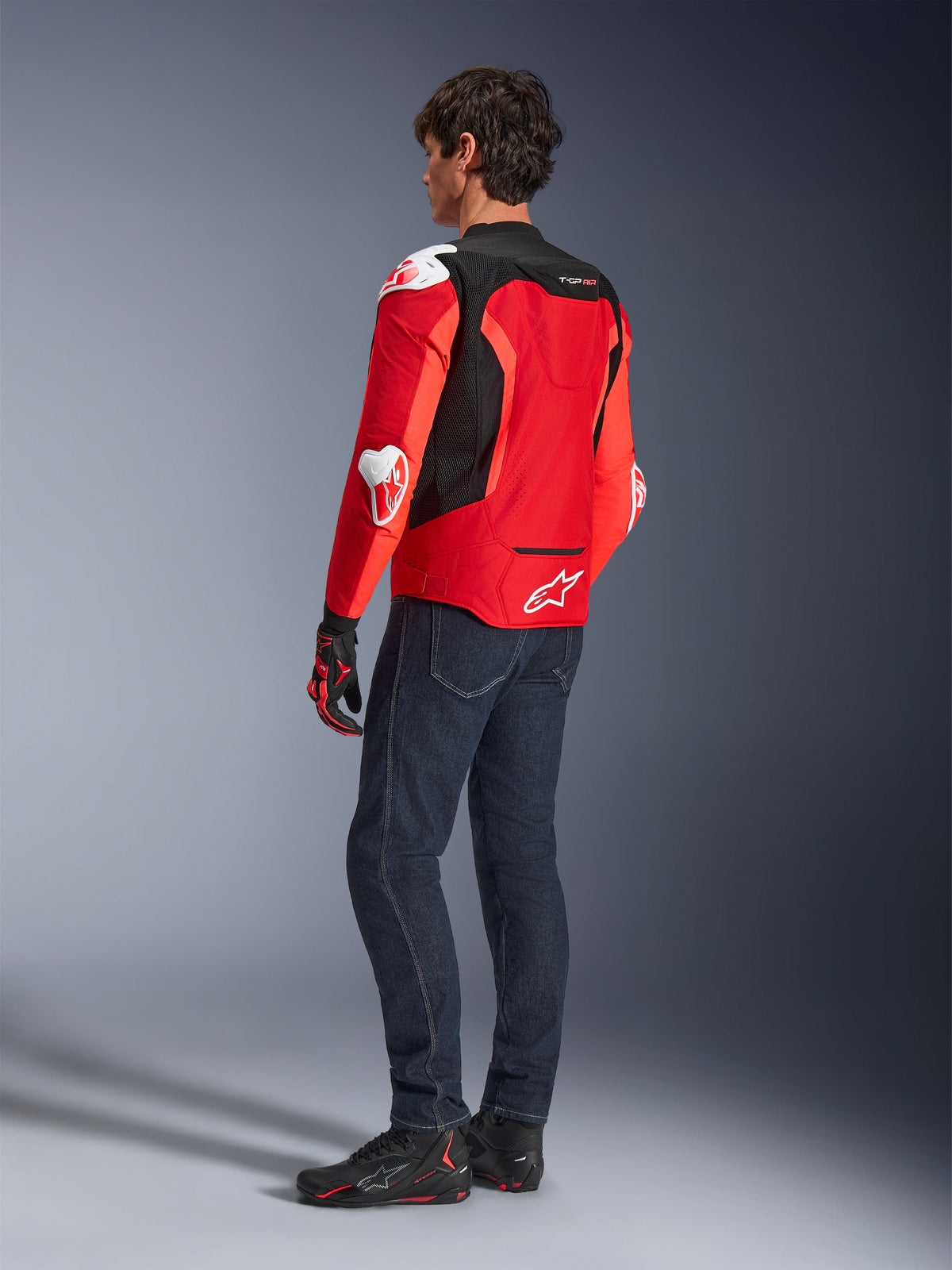 Casaco Alpinestars T-GP Air, vista traseira de um homem a usar um casaco têxtil em vermelho vivo e vermelho fluorescente com painéis de malha preta e sliders de proteção brancos, combinado com calças de ganga azul-escuro e sapatos de condução pretos, equipamento de motociclismo desportivo