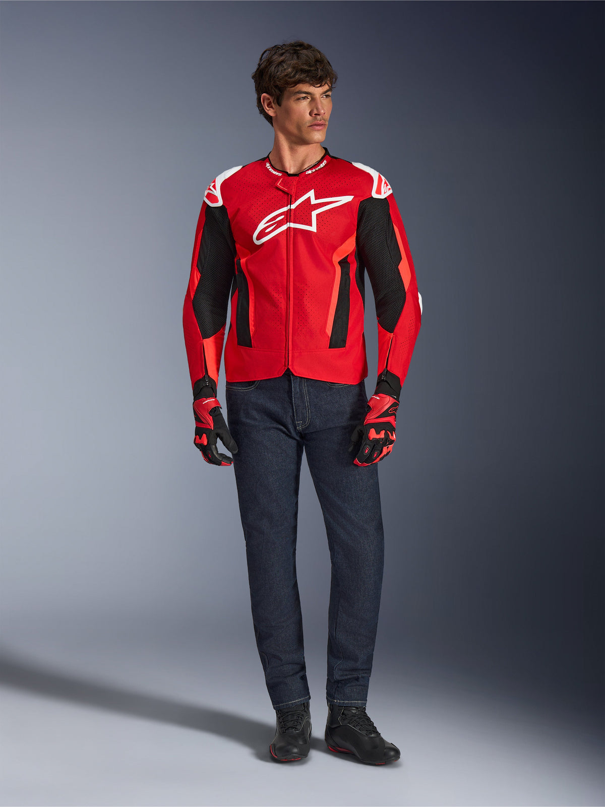 Casaco Alpinestars T-GP Air, Vermelho Brilhante, Vermelho Fluo e Preto, modelo masculino a usar equipamento completo de motociclismo com calças de ganga azul-escuro e luvas a condizer, casaco têxtil desportivo com painéis de malha perfurada e logótipo Astars branco, fotografia de estúdio.