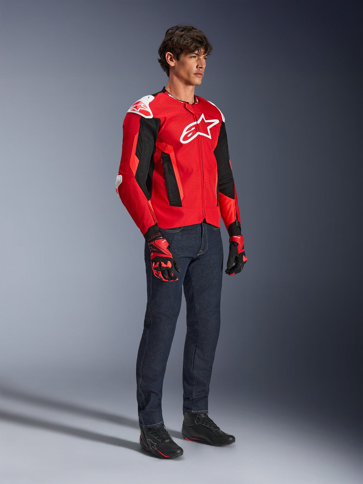Casaco Alpinestars T-GP Air, casaco desportivo de motociclismo, vermelho brilhante e preto com detalhes em branco, usado por um modelo com calças de ganga azul-escuro e luvas de corrida vermelhas, com painéis de malha de alto fluxo e sliders de ombro brancos, ajuste desportivo aerodinâmico para condução em clima quente.