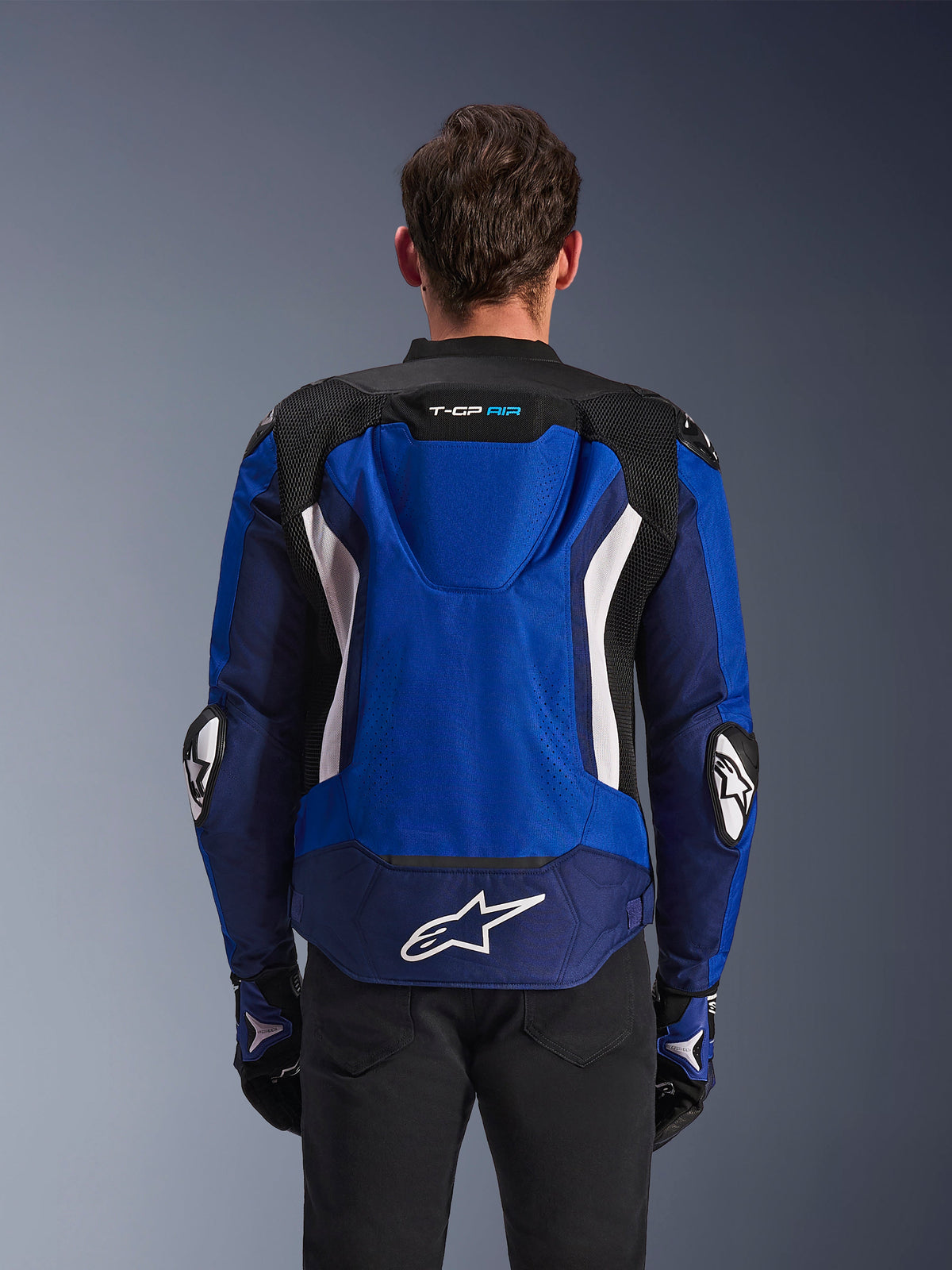 Alpinestars T-GP Air Jacket, Sport Textile Motorcycle Jacket, Bright Blue Navy and White, rear view featuring the integrated aerodynamic hump and large white logo on lower back, worn with matching blue and black riding gloves