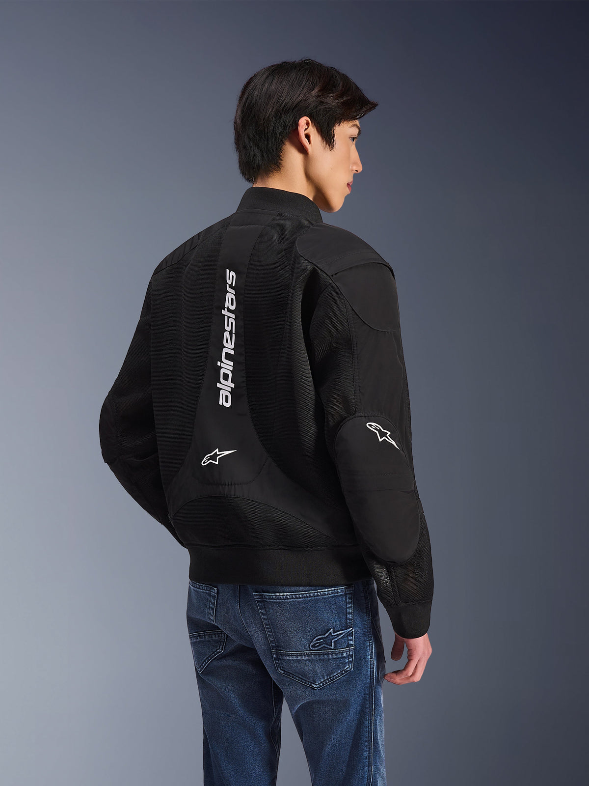 Casaco Alpinestars Flight Air, casaco de motociclismo preto, vista traseira com logótipo branco vertical grande e painéis de ventilação em malha, estilo bomber com bainha canelada, usado com calças de ganga azuis, equipamento de condução urbano contemporâneo