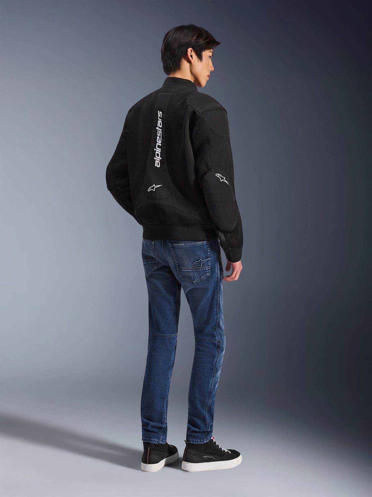 Modelo a usar o Casaco Alpinestars Flight Air em preto, visto de costas, combinado com calças de ganga de motociclismo azuis e sapatilhas pretas, com a marca Alpinestars vertical em branco e painéis de ventilação em malha 3D.