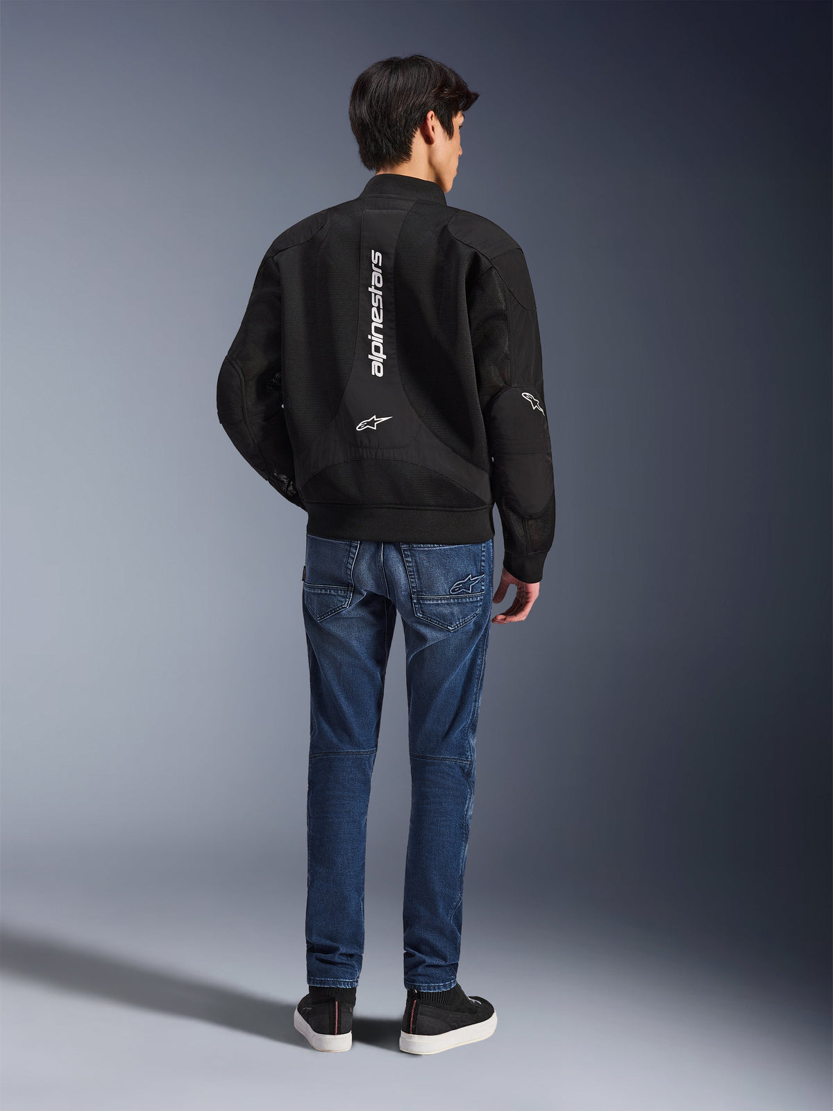 Modelo a usar o casaco Alpinestars Flight Air visto de costas, casaco de motociclismo preto estilo bomber com painéis de malha 3D e logótipo vertical branco, combinado com calças de ganga azuis e sapatilhas pretas, equipamento de motociclismo urbano contemporâneo