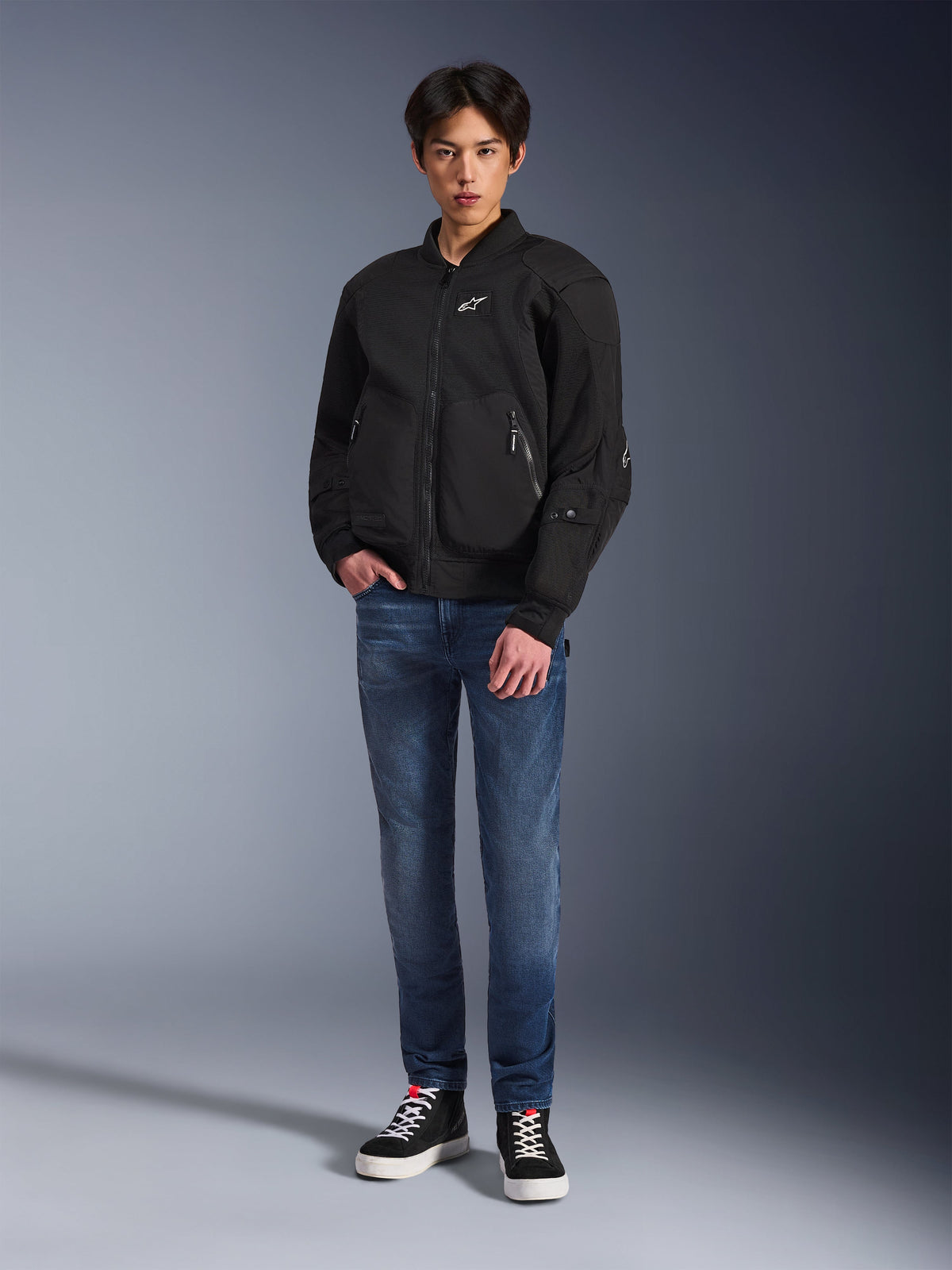 Modelo a usar o casaco Alpinestars Flight Air em preto, um casaco de motociclismo urbano de estilo bomber com painéis de malha 3D e fecho frontal, combinado com calças de ganga azuis e sapatilhas pretas, vestuário de condução contemporâneo de inspiração urbana.