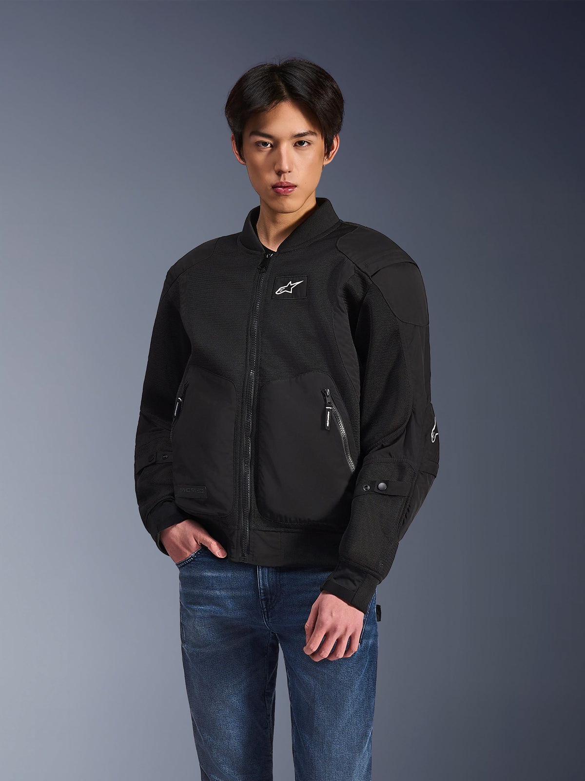 Casaco Alpinestars Flight Air, casaco de motociclismo urbano preto, usado por um homem com calças de ganga azuis, com uma estética de estilo bomber, painéis de malha, fecho de correr frontal e logótipo branco no peito, concebido para condução urbana em tempo quente