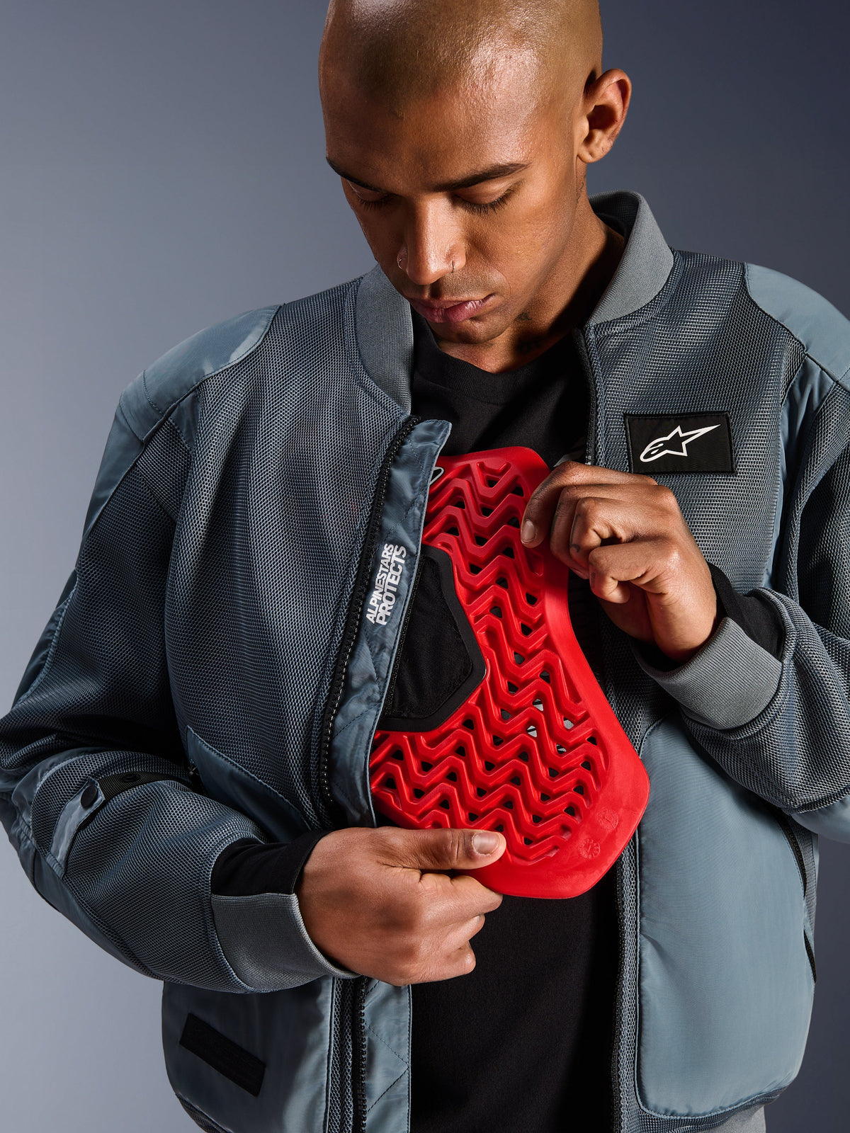 Casaco Alpinestars Flight Air em Cinzento Storm, casaco bomber de motociclismo urbano com painéis em malha 3D para circulação de ar, aberto para revelar uma inserção de protetor de costas Nucleon vermelho, com a marca 'Alpinestars Protects' e uma estética streetwear moderna