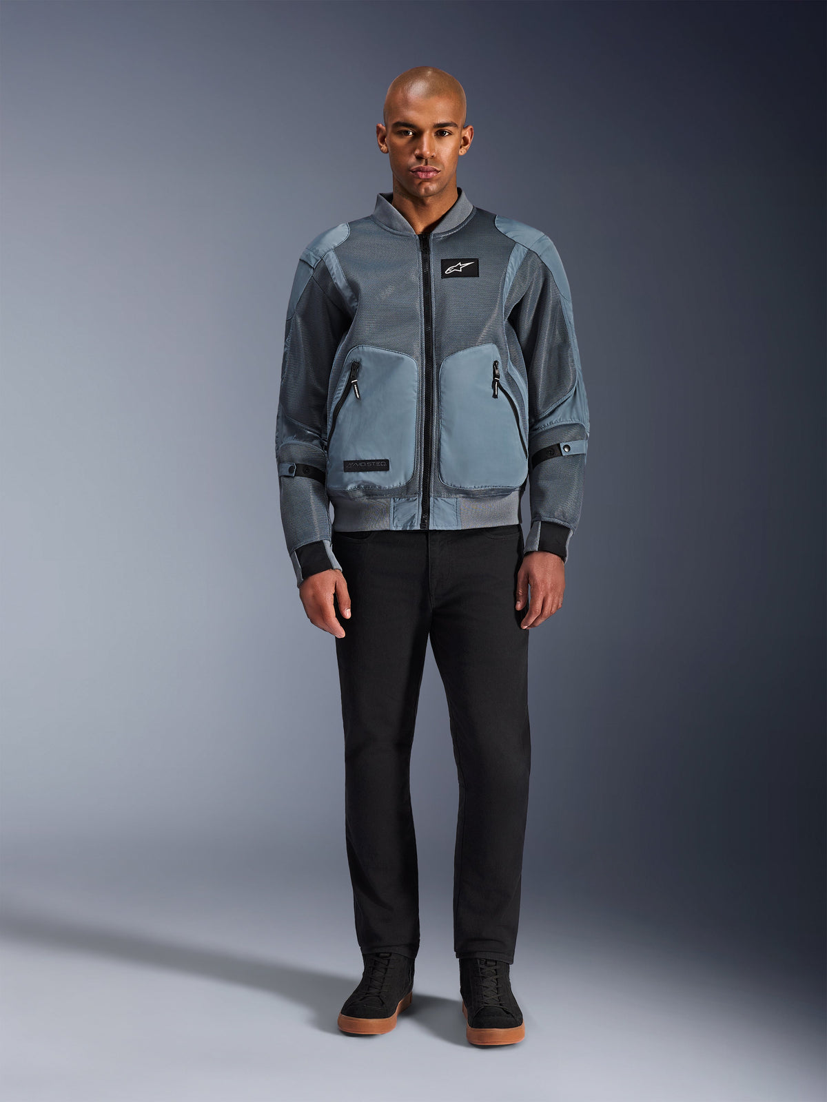 Casaco Alpinestars Flight Air em Storm Gray, casaco de motociclismo urbano inspirado no estilo bomber com painéis de malha 3D e bolsos frontais, vestido por um homem com calças pretas e ténis de cano alto pretos, vestuário de condução moderno de estilo urbano