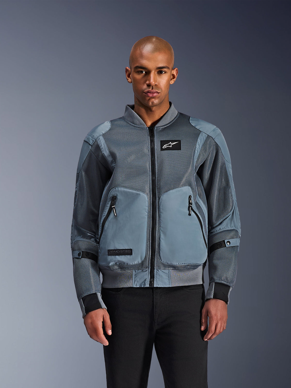 Casaco Alpinestars Flight Air, casaco de motociclismo estilo bomber urbano, cinzento Storm Gray, com painéis de ventilação em malha 3D e emblema do logótipo Astars branco, usado por modelo masculino com calças pretas, vestuário contemporâneo de condução urbana