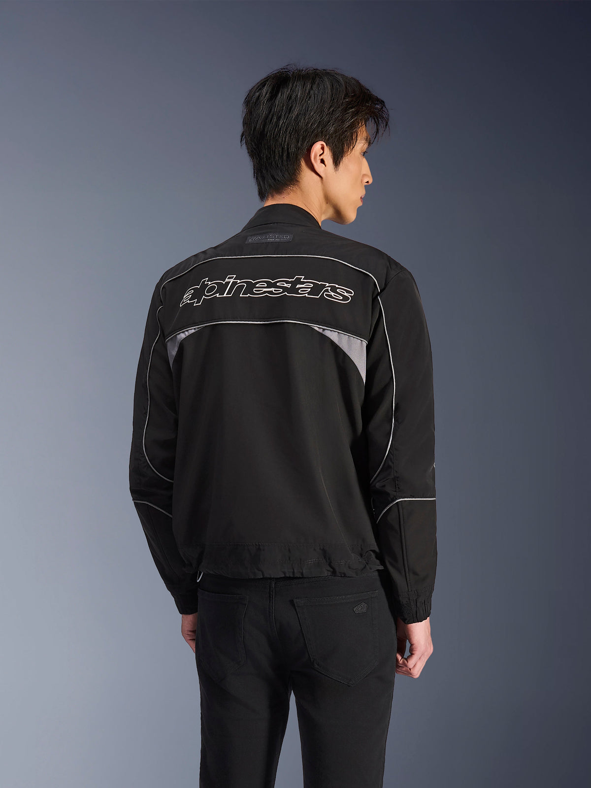 Casaco Alpinestars Unite, casaco de motociclismo urbano, preto com detalhes cinzento escuro, vista traseira com logótipo Alpinestars branco nos ombros, debrum e painéis refletores, design minimalista de streetwear em softshell