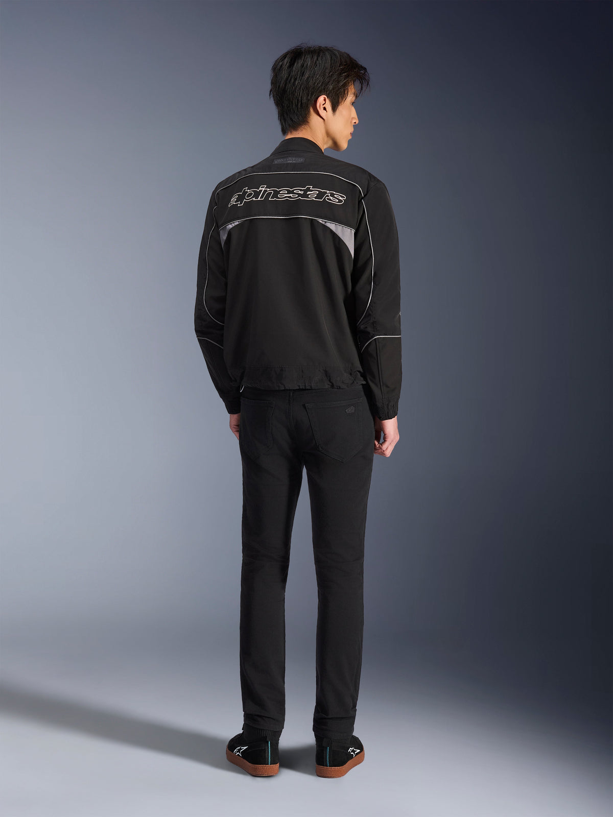 Alpinestars Unite Jacket, casaco de motociclismo preto e cinzento escuro, vista traseira mostrando o logótipo Alpinestars em contorno nas costas, painéis refletores cinzentos, design urbano minimalista, usado com calças pretas e sapatos pretos