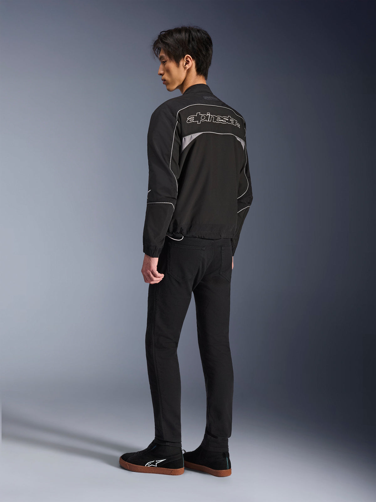 Casaco Alpinestars Unite, casaco urbano de motociclismo, preto e cinzento escuro, vista traseira com logótipo Alpinestars grande e contornado, debrum e painéis refletores, design softshell minimalista, usado com calças pretas e calçado casual