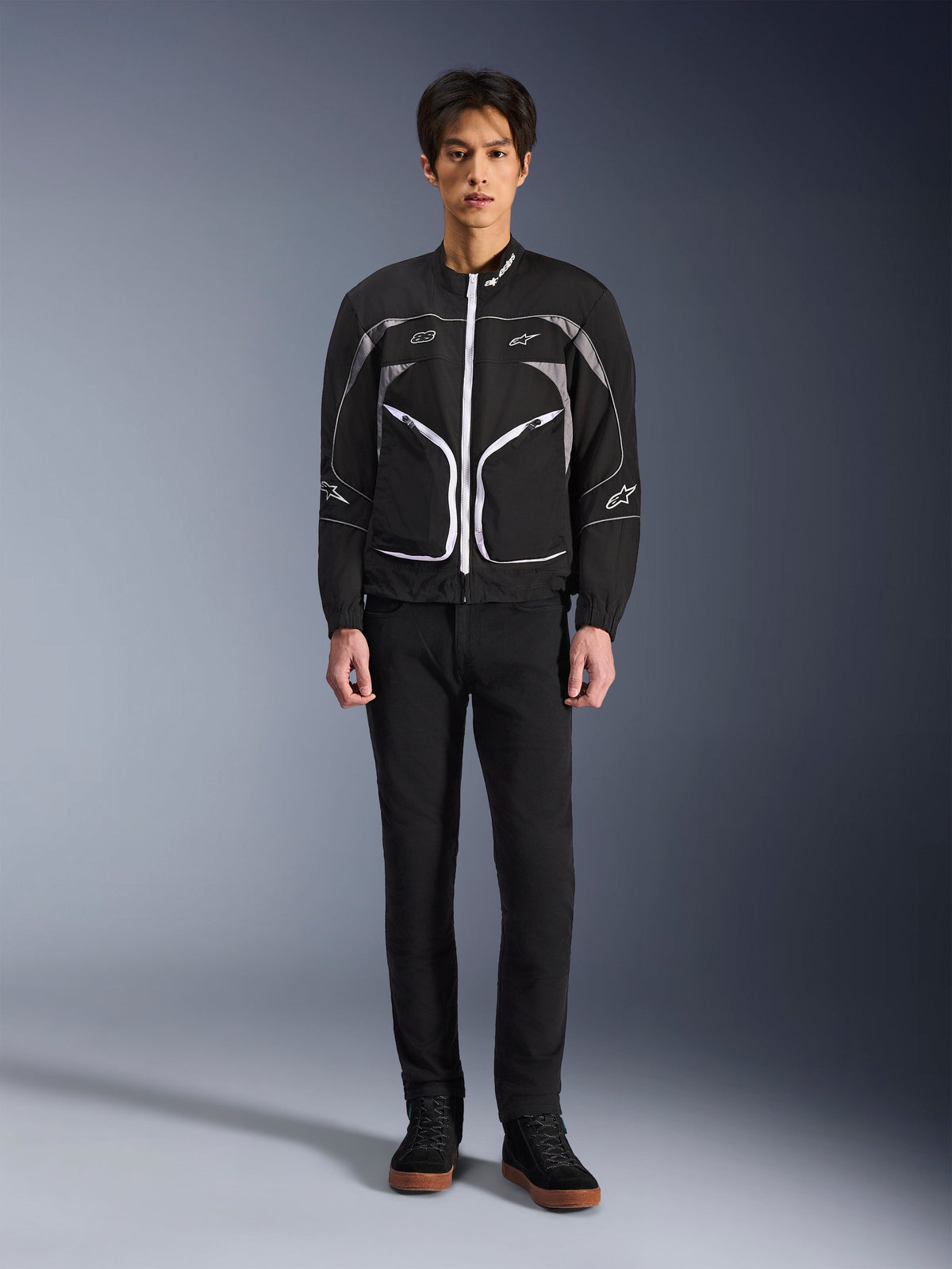 Casaco Alpinestars Unite, casaco de motociclismo, preto e cinzento escuro, usado por um modelo com calças pretas e botas pretas, com fechos de correr brancos em contraste, debrum refletor e design urbano minimalista
