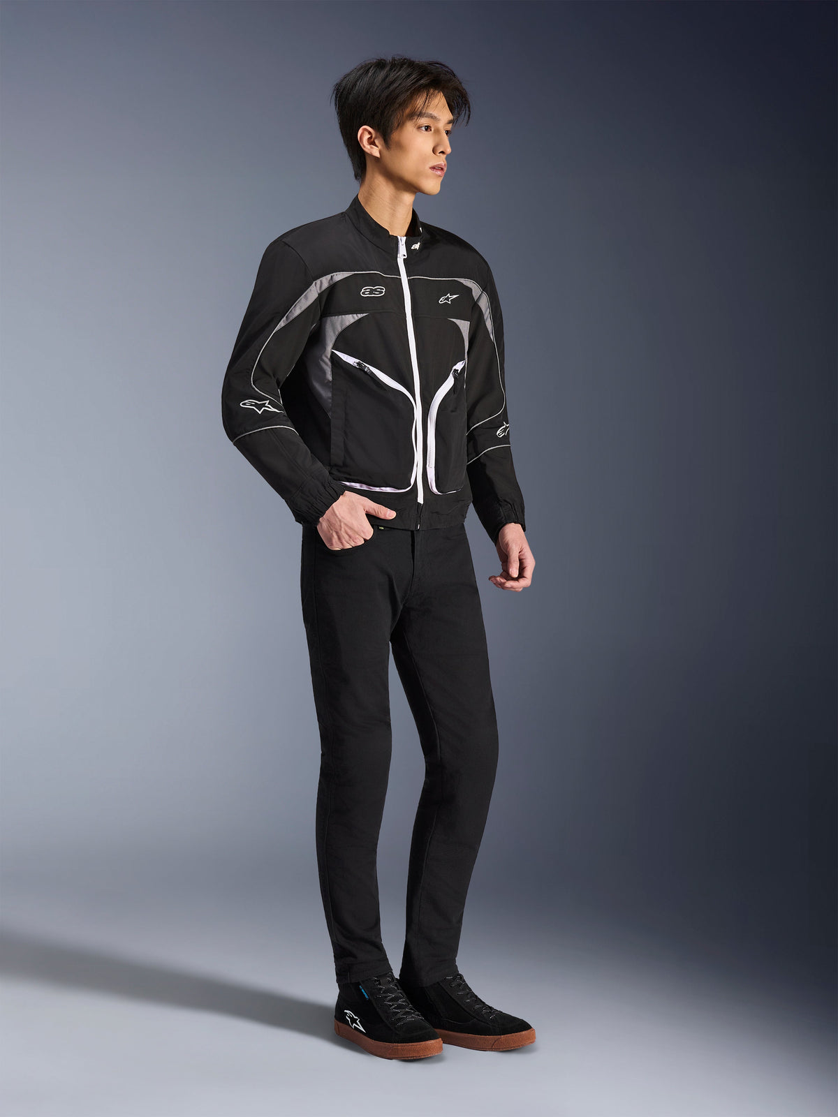 Modelo a usar o Casaco Unite da Alpinestars em preto e cinzento escuro, casaco de motociclismo urbano com fechos de correr brancos e detalhes refletores, combinado com calças pretas slim-fit e sapatilhas, equipamento de condução minimalista de inspiração urbana