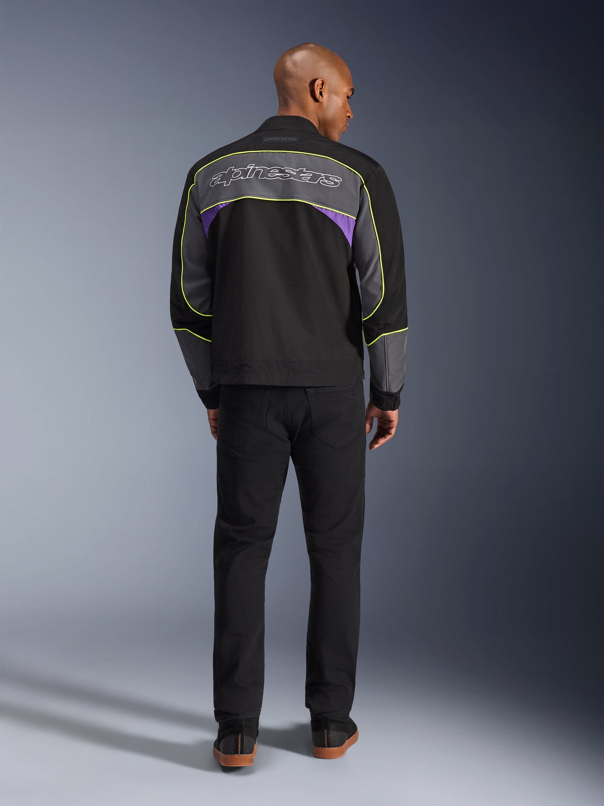 Casaco Alpinestars Unite, casaco de motociclismo urbano em Black Asphalt Violet, vista traseira com painéis cinzentos, detalhes roxos e vivos em amarelo néon, logótipo Alpinestars em letra cursiva nos ombros, usado com calças pretas