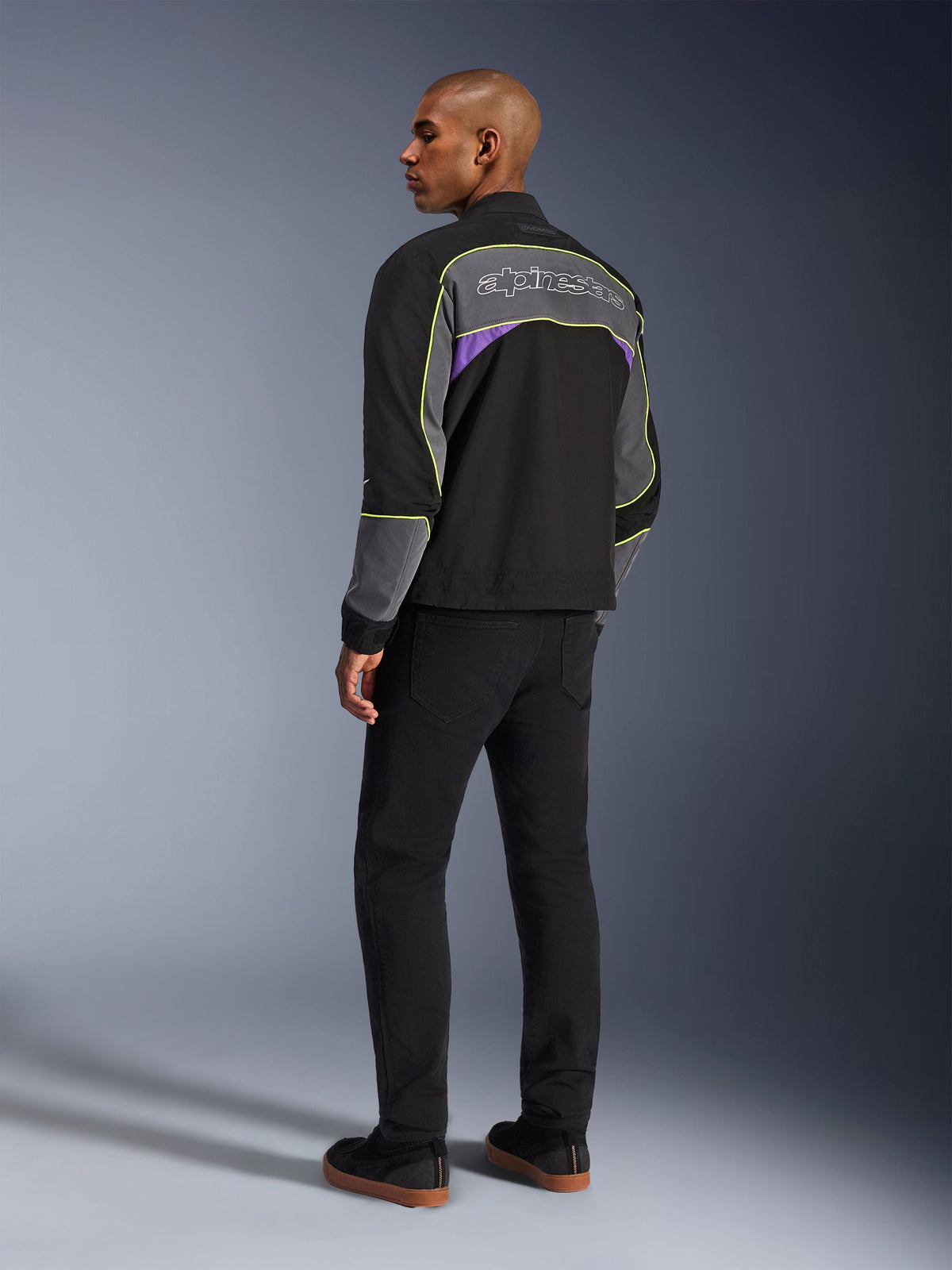 Casaco Alpinestars Unite, casaco de motociclismo urbano, Black Asphalt Violet, vista traseira de três quartos num modelo, softshell preto com painéis cinzentos e pormenores violeta, debrum refletor amarelo néon, logótipo Alpinestars na parte superior das costas, combinado com calças pretas e sapatos casuais pretos