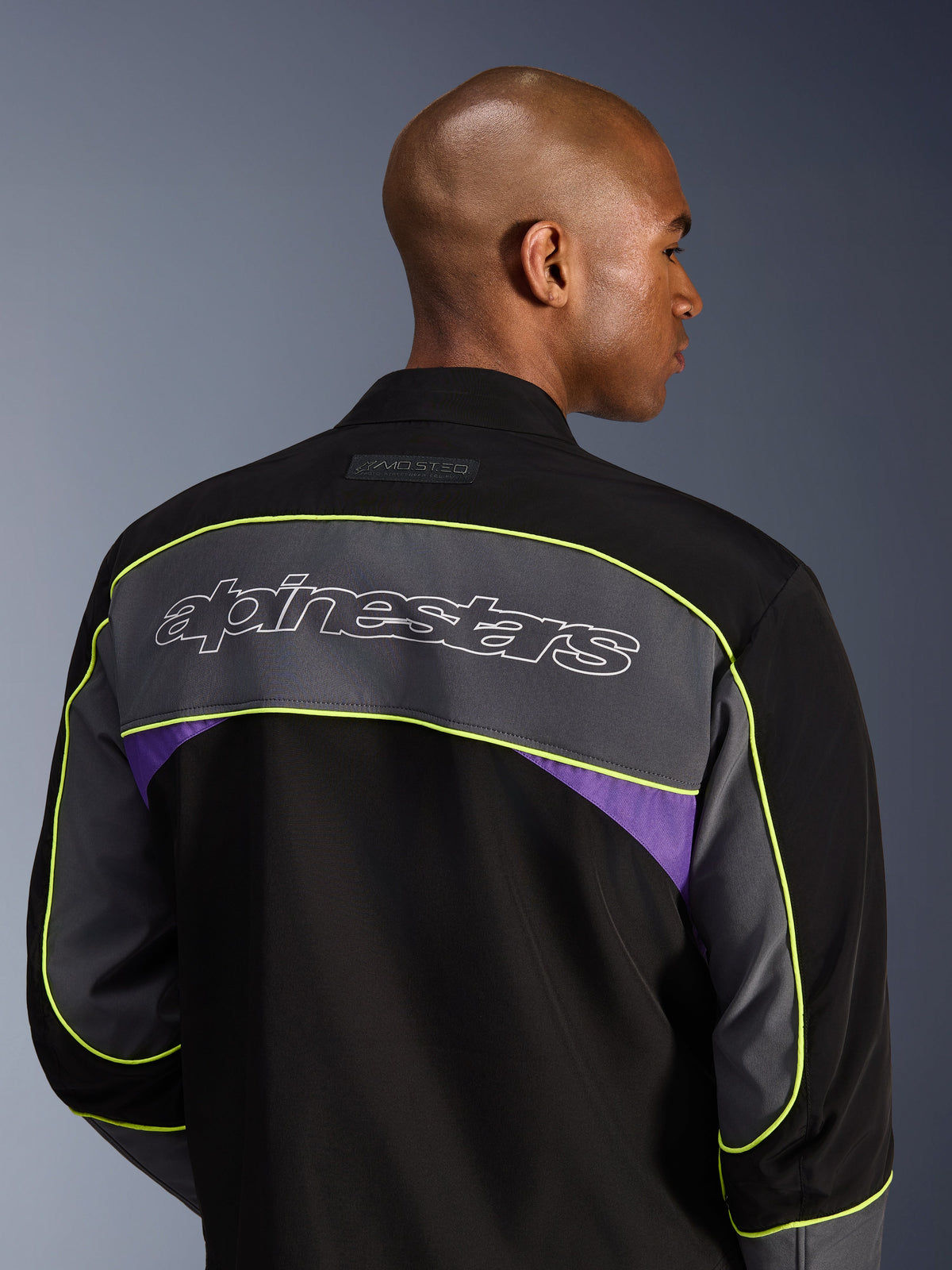 Casaco Alpinestars Unite, casaco de motociclismo urbano, Black Asphalt Violet, vista traseira que mostra um grande painel cinzento nos ombros com o logótipo Alpinestars, debruado a amarelo néon e detalhes em violeta numa construção softshell de 3 camadas
