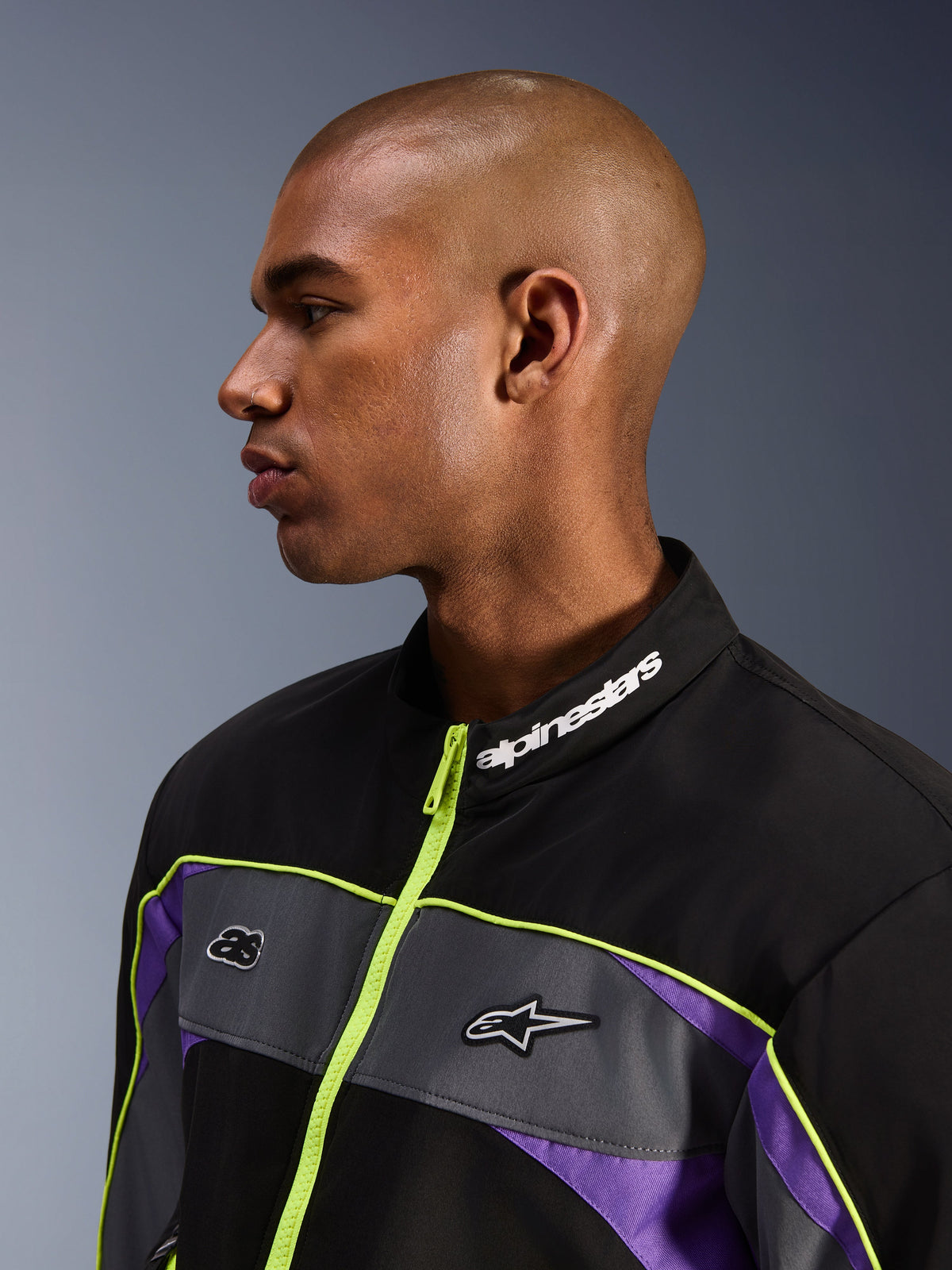 Casaco Alpinestars Unite, Black Asphalt Violet, vista de perfil de modelo masculino com foco na gola alta e marca Alpinestars em branco, fecho de correr amarelo néon, painéis cinzentos no peito com logótipos, detalhes roxos, estilo minimalista de streetwear urbano para motociclismo