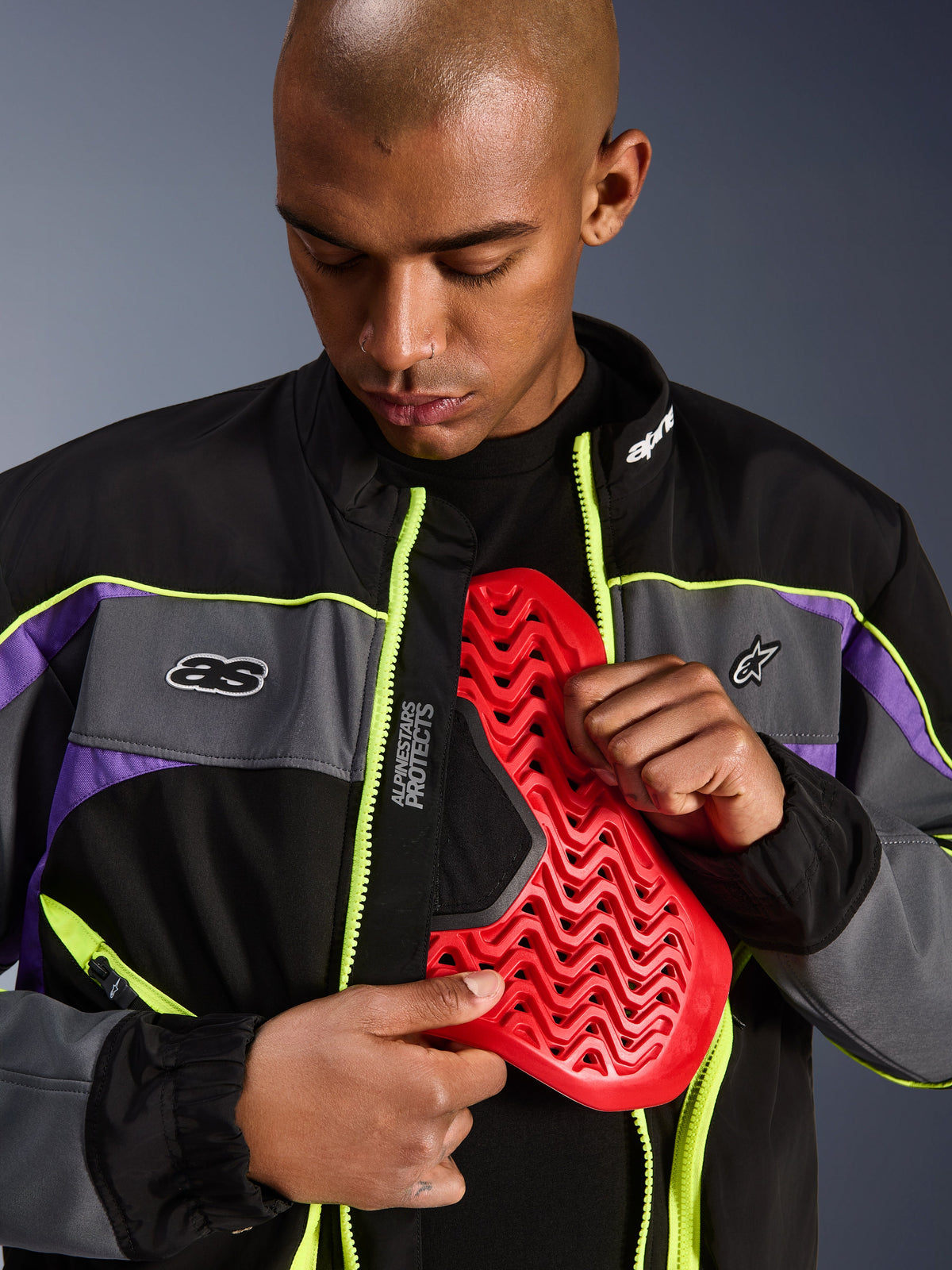 Casaco Alpinestars Unite, casaco de motociclismo urbano, preto, asfalto e violeta com detalhes em amarelo néon, modelo masculino a mostrar uma proteção de peito Nucleon vermelha no interior do casaco aberto, estética streetwear minimalista para condução urbana