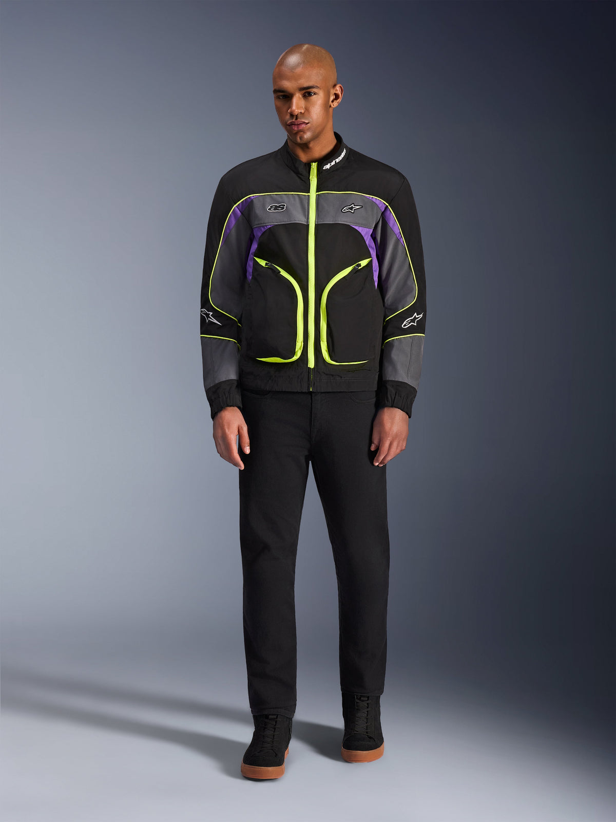 Modelo masculino a usar o Casaco Alpinestars Unite em Black Asphalt Violet, com base preta, painéis cinzentos e roxos, fechos e acabamentos em amarelo néon vibrante, combinado com calças e sapatos pretos, casaco de motociclismo urbano minimalista