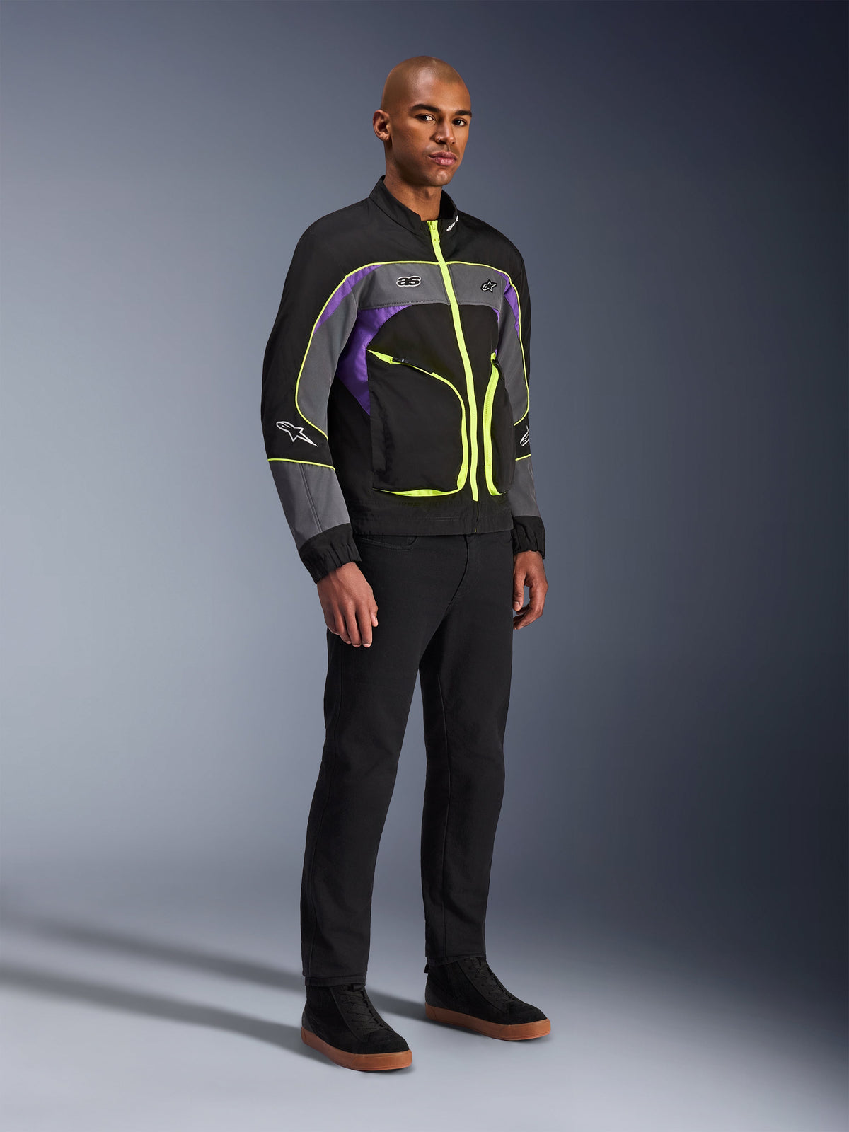 Casaco Alpinestars Unite, casaco de motociclismo urbano, Black Asphalt Violet com detalhes em amarelo néon, vestido por um modelo com calças pretas e sapatilhas, estilo streetwear contemporâneo para condução urbana