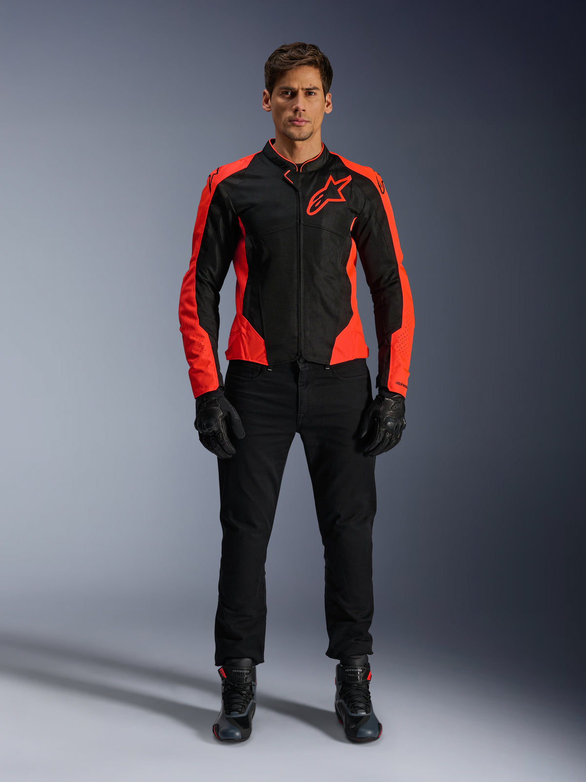 Modelo masculino a usar o Casaco Alpinestars Viper Air V4 em Vermelho Fluo e Preto, combinado com calças de motociclismo, luvas e botas pretas, apresentando painéis de malha respirável para ventilação e um logótipo de estrela proeminente no peito, vestuário desportivo para motociclismo de estrada