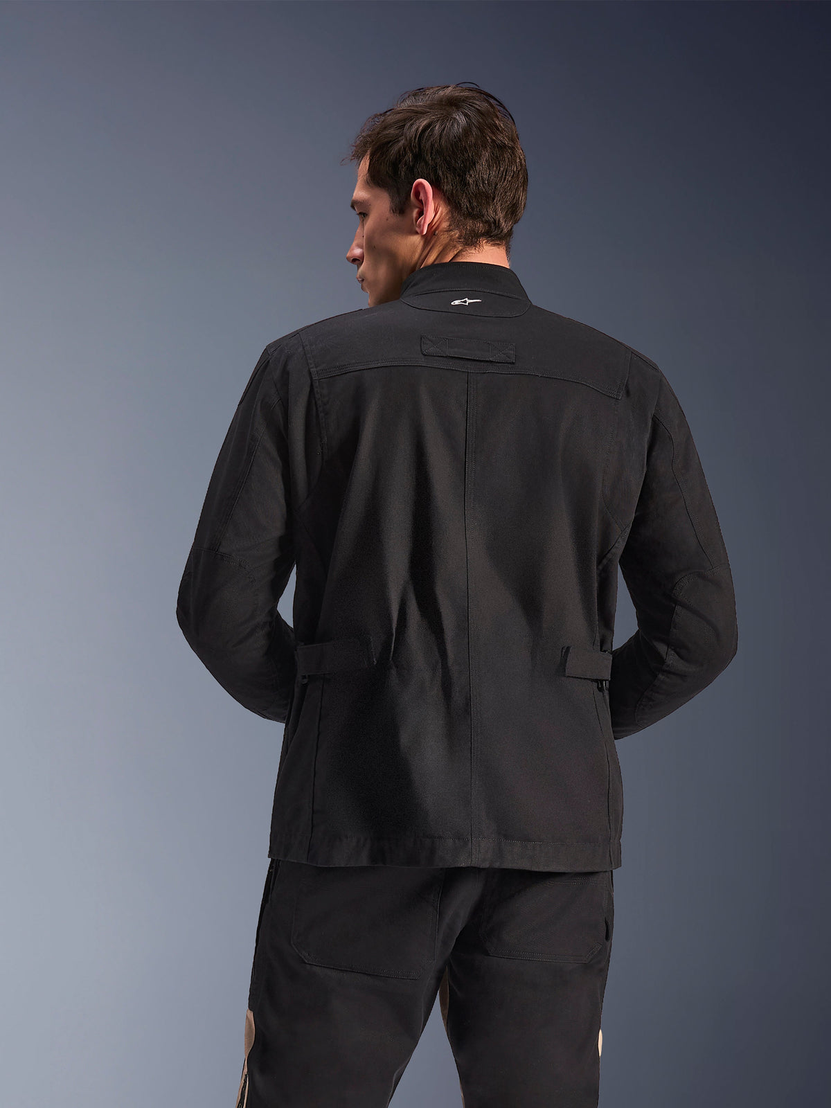 Alpinestars Dice Jacket, casaco de motociclista urbano, preto, vista traseira mostrando ombros pregueados e abas de ajuste na cintura, estética de casaco de campo com pequeno logótipo branco no colarinho, equipamento têxtil de proteção para condução urbana