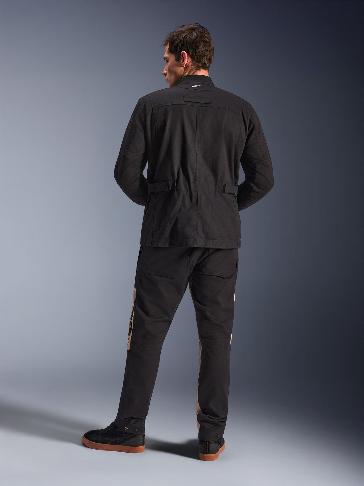 Vista traseira do Casaco Alpinestars Dice em Preto, casaco de campo urbano para motociclismo com ombros pregueados e ajustadores de cintura, combinado com calças de condução pretas e beges e sapatilhas, estilo citadino contemporâneo