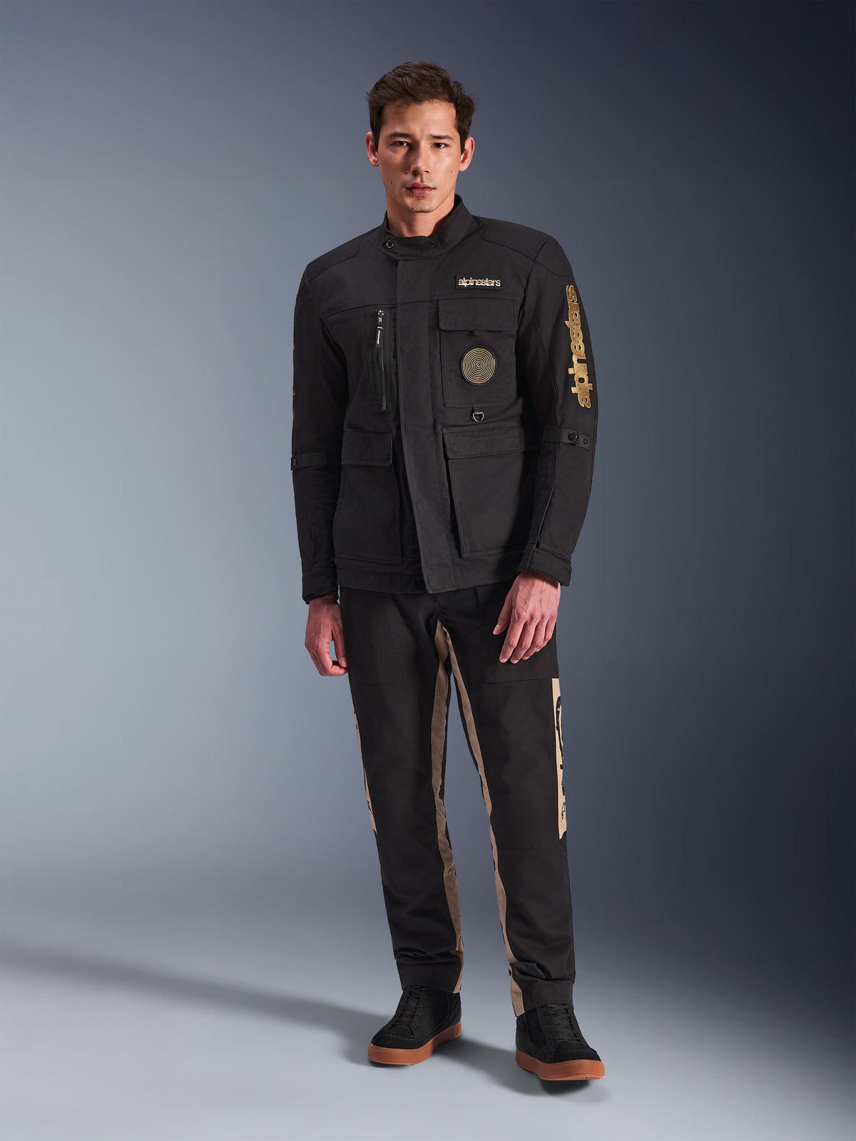 Modelo masculino a usar o Casaco Dice da Alpinestars, casaco de motociclista urbano preto com estilo vintage de casaco de campo, quatro bolsos frontais funcionais, marca Alpinestars na manga e no peito, combinado com calças pretas e beges, equipamento de condução urbana moderno