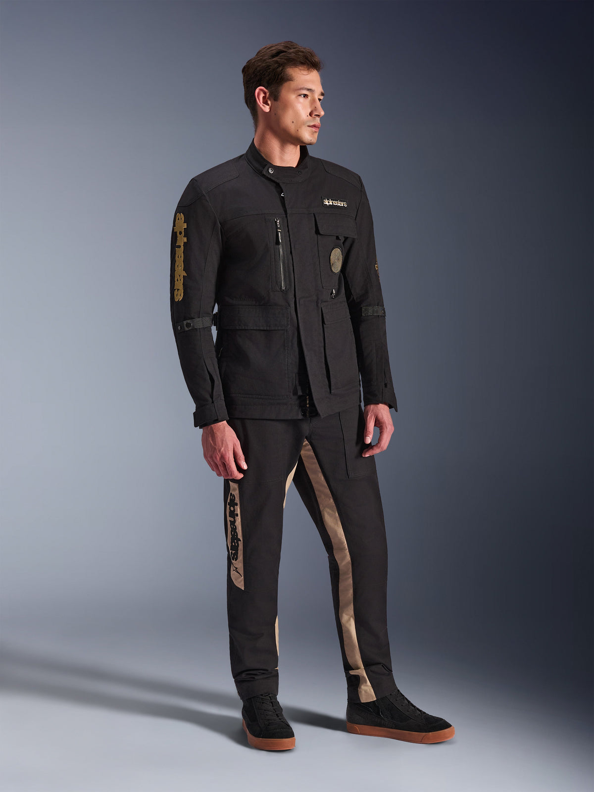 Modelo masculino a usar o Casaco Alpinestars Dice em preto, casaco de motociclista urbano com estética vintage de casaco de campo, com múltiplos bolsos utilitários e logótipo dourado na manga, combinado com calças pretas e beges a condizer e sapatilhas pretas