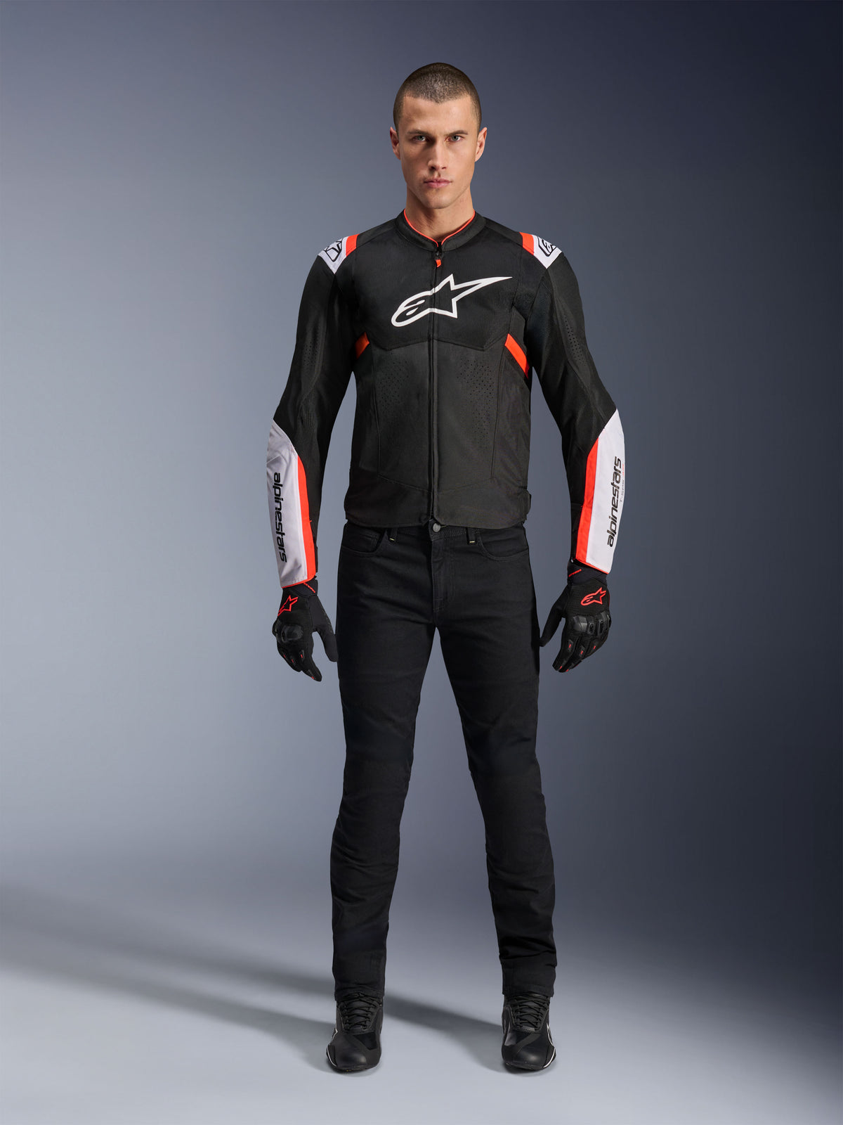 Modelo masculino a usar o Casaco Alpinestars T-Sps Air V2 em Preto, Branco e Vermelho Fluo, com grandes painéis de malha técnica e logótipos brancos no peito e mangas, calças e luvas técnicas pretas, equipamento de motociclismo urbano desportivo