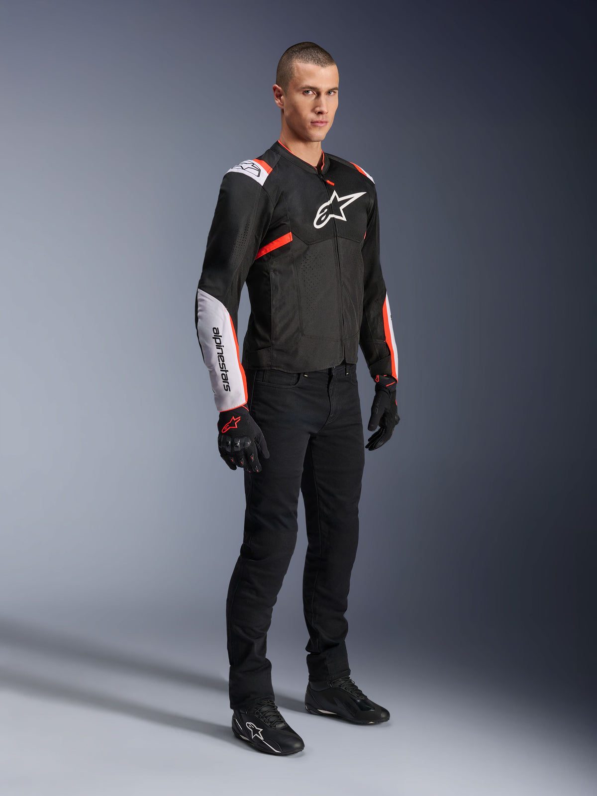 Casaco Alpinestars T-Sps Air V2, preto, branco e vermelho fluo, usado por um modelo masculino com calças e luvas de motociclismo pretas, casaco têxtil com grandes painéis de malha, secções brancas nos ombros e antebraços, e logótipo Alpinestars no peito