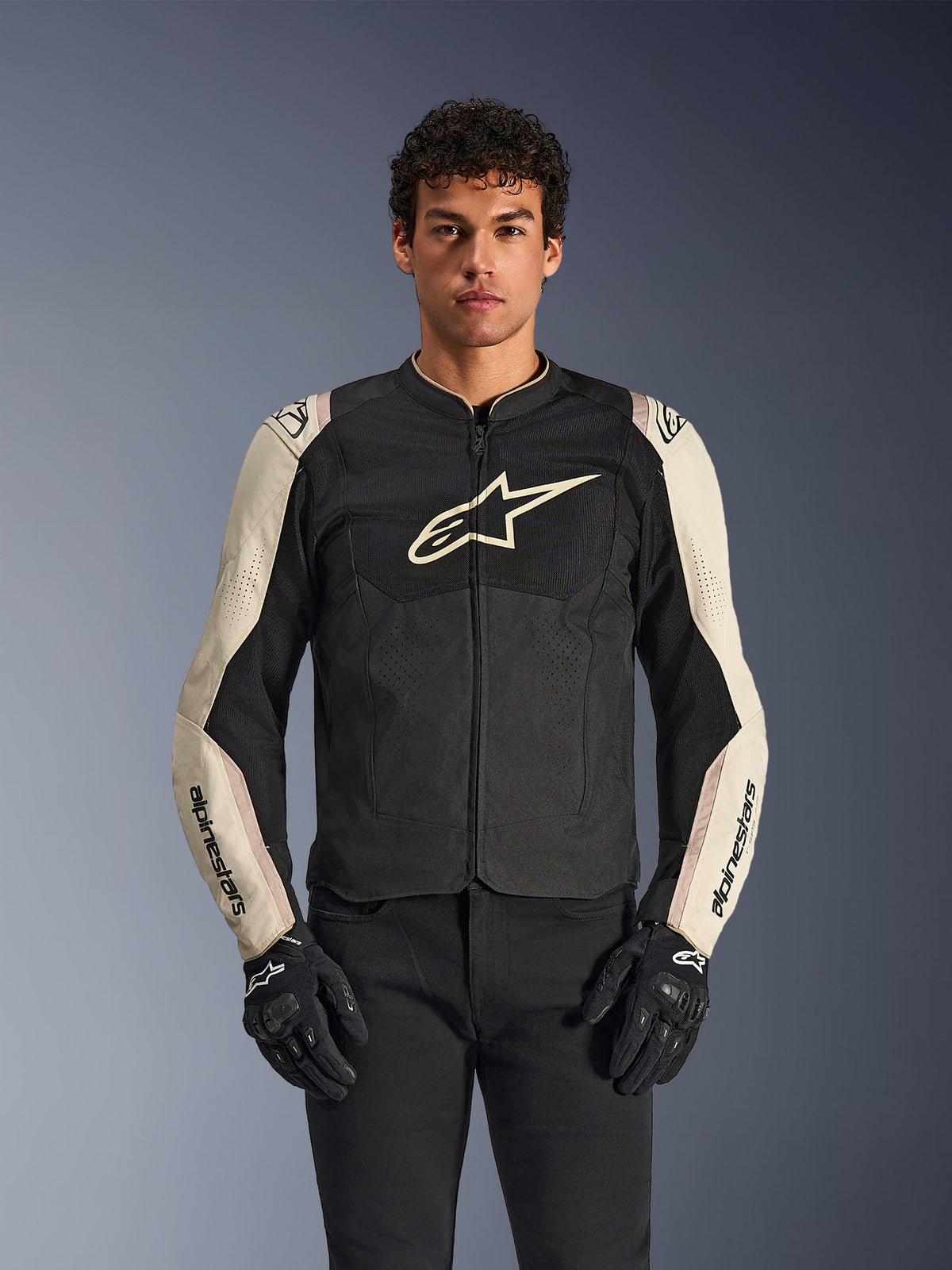 Casaco Alpinestars T-SPS Air V2, casaco de motociclismo, preto e alumínio mastic, modelo masculino a usar o casaco com calças e luvas pretas, com painéis de malha para ventilação, logótipo Alpinestars de grandes dimensões no peito e design desportivo ajustado