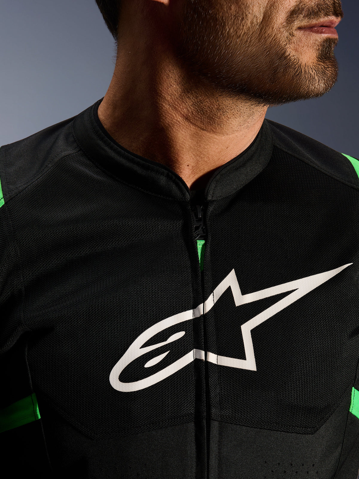 Pormenor do Casaco Alpinestars T-SPS Air V2, Preto e Verde Fluo, com painéis de malha técnica respirável, fecho central e o proeminente logótipo de estrela da Alpinestars em branco no peito, equipamento de motociclismo de alta performance