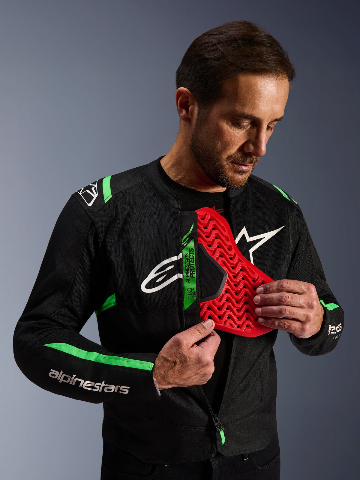 Casaco Alpinestars T-SPS Air V2, casaco de motociclismo preto e verde flúor, homem a demonstrar a inserção de protetor de peito Nucleon vermelho, etiqueta Tech-Air Ready, design têxtil desportivo com painéis de malha técnica