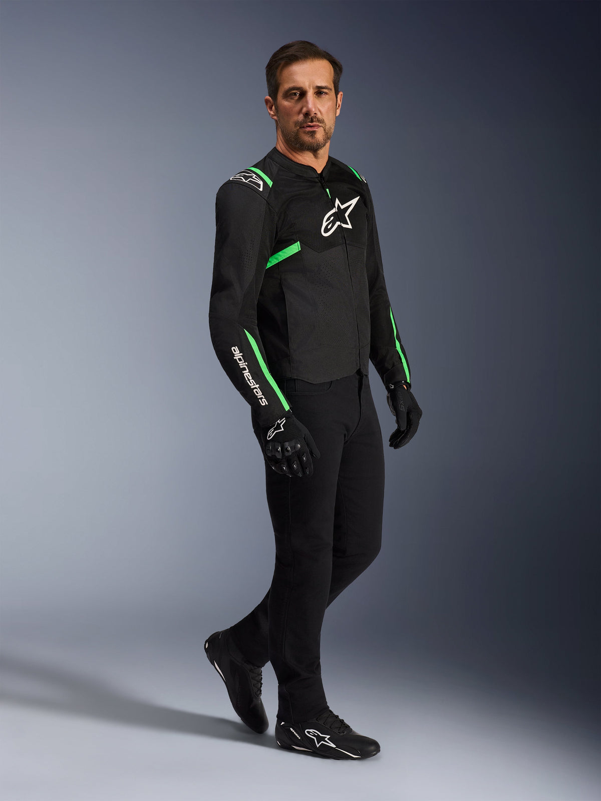 Modelo masculino a usar o Casaco Alpinestars T-SPS Air V2 em preto e verde néon, combinado com calças de condução e luvas pretas, casaco têxtil de corte desportivo com painéis de malha e detalhes néon, equipamento de motociclismo urbano completo