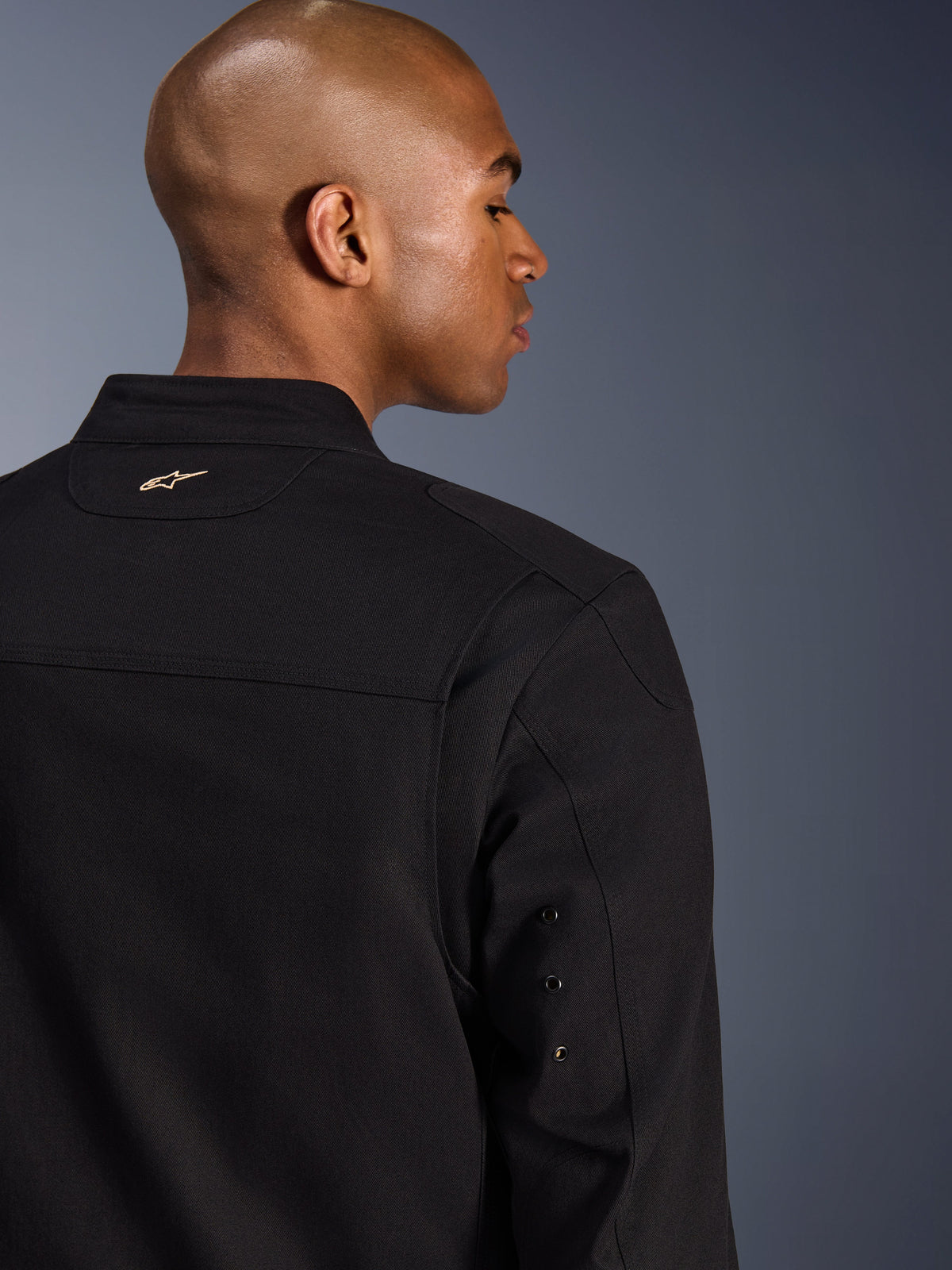 Alpinestars Bruiser Jacket, textile urban motorcycle jacket, Black, close-up back view featuring an embroidered gold logo on the upper back, metal eyelet sleeve details, and pleated shoulder panels for enhanced mobility.