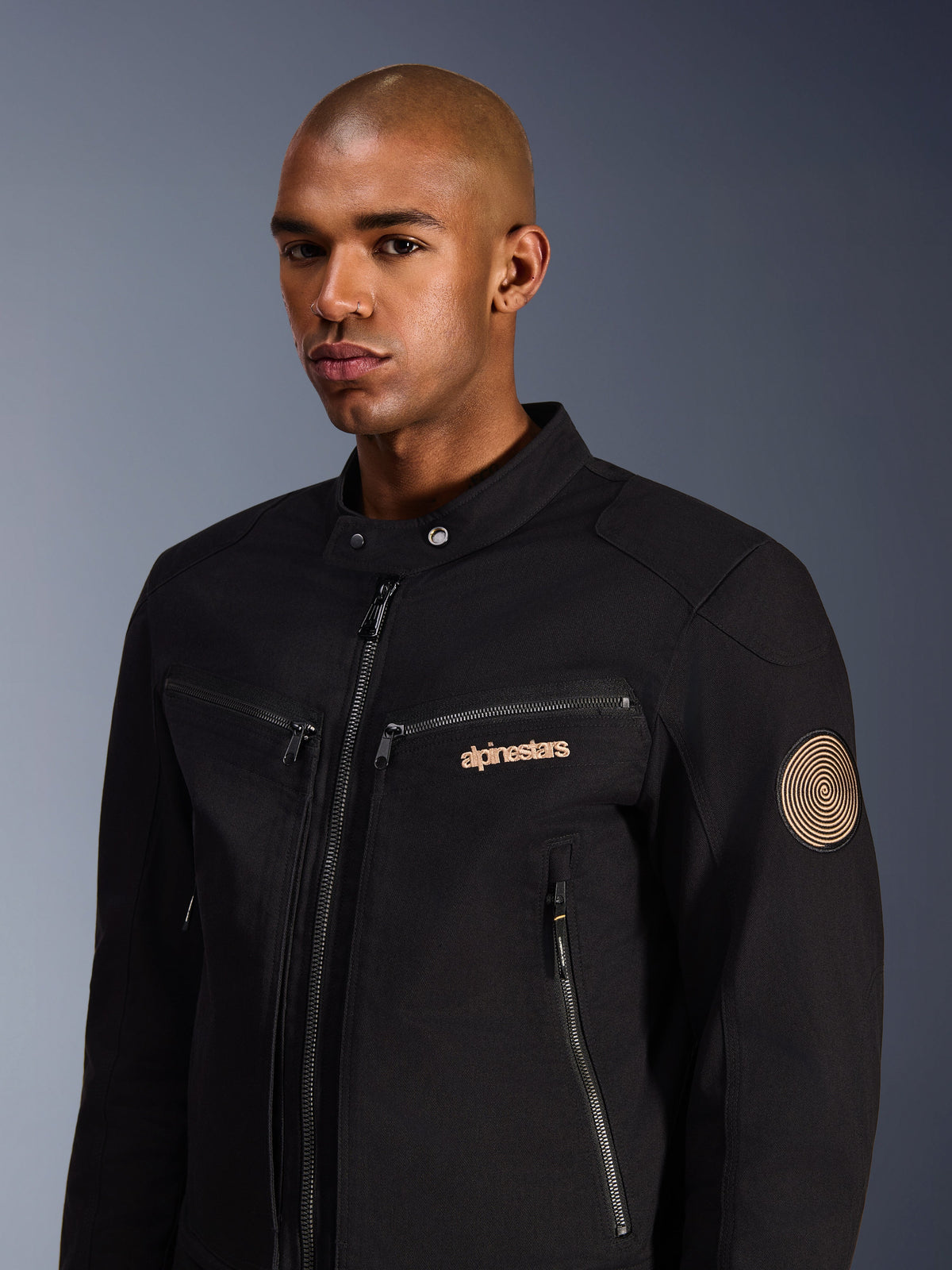 Male model wearing Alpinestars Bruiser Jacket, Black, urban motorcycle jacket in polycotton twill, featuring zippered pockets, gold embroidered logo on the chest, and Oscar spiral patch on the sleeve, modern commuting style
