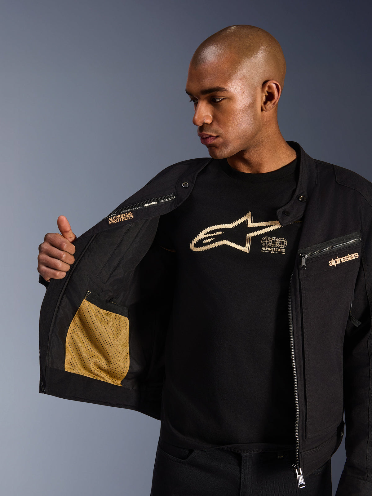 Alpinestars Bruiser Jacket, Black urban motorcycle jacket, close-up view showing open interior with gold mesh pocket and 'Alpinestars Protects' branding, worn over a black logo t-shirt, textile riding gear with workwear aesthetic