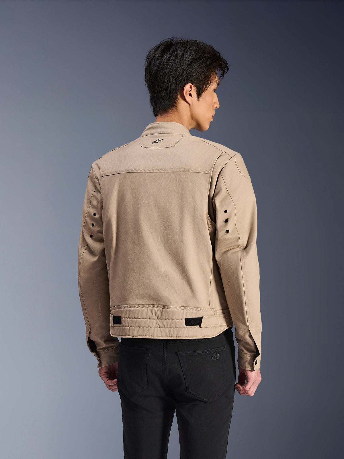Casaco Alpinestars Bruiser, casaco de motociclismo urbano castanho, vista traseira com ombros articulados com pregas e tiras de ajuste na cintura, usado com calças pretas, equipamento de condução contemporâneo inspirado no vestuário de trabalho