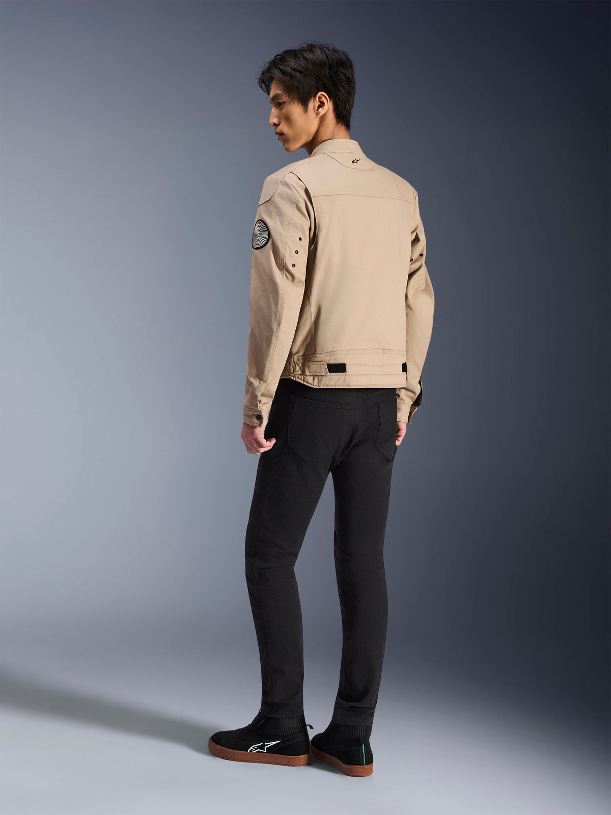 Modelo masculino visto de costas a usar o casaco Alpinestars Bruiser Jacket em bege, um casaco de motociclismo urbano contemporâneo com estilo inspirado em vestuário de trabalho, apresentando um patch circular na manga, tiras de ajuste na cintura e ombros articulados, combinado com calças pretas e ténis pretos.