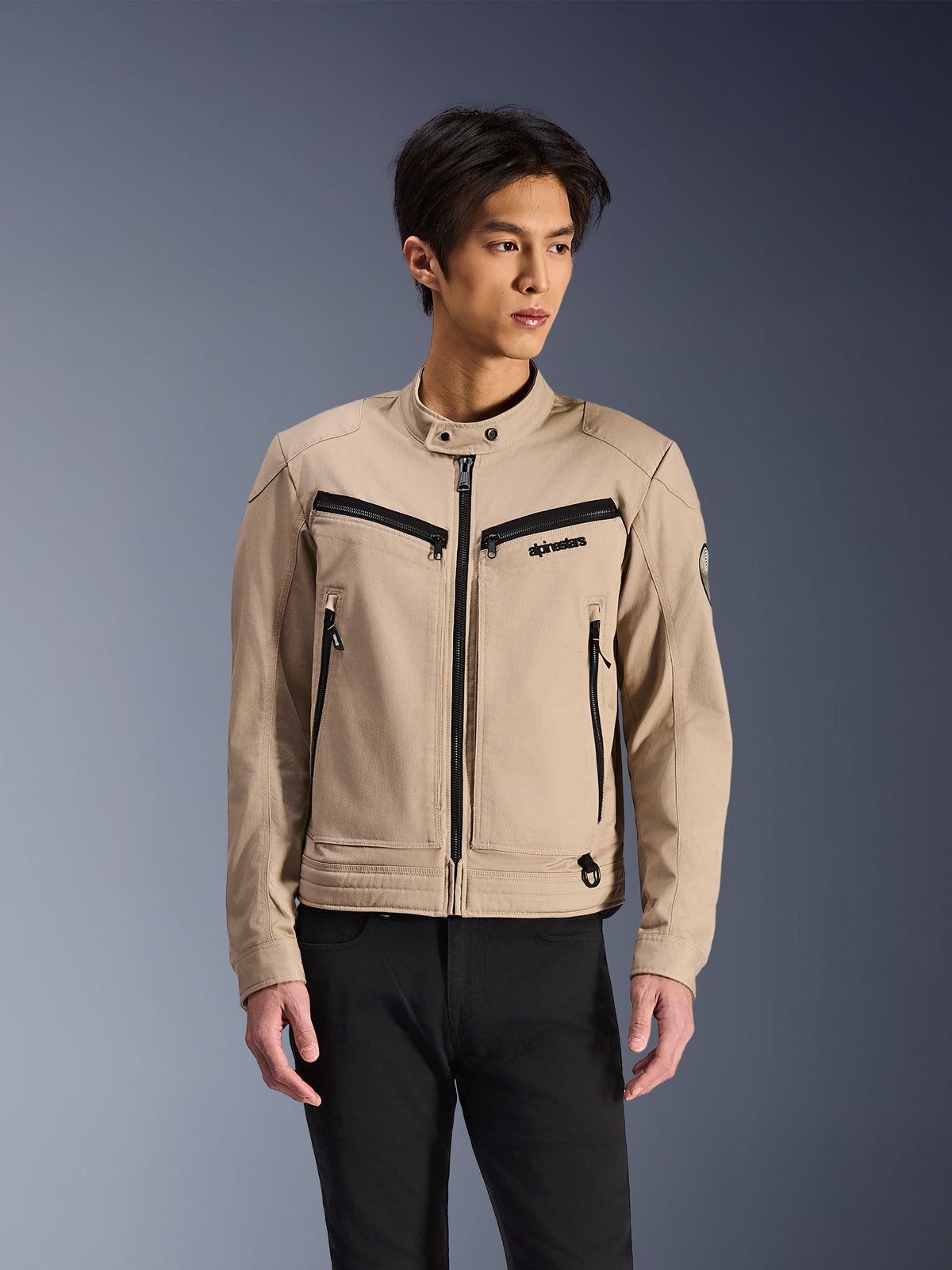 Alpinestars Bruiser Jacket, casaco de motociclismo bege usado por um modelo masculino, com detalhes de fechos pretos no peito e bolsos laterais, gola alta com botões de pressão, vestuário de condução urbana inspirado em roupa de trabalho, combinado com calças pretas
