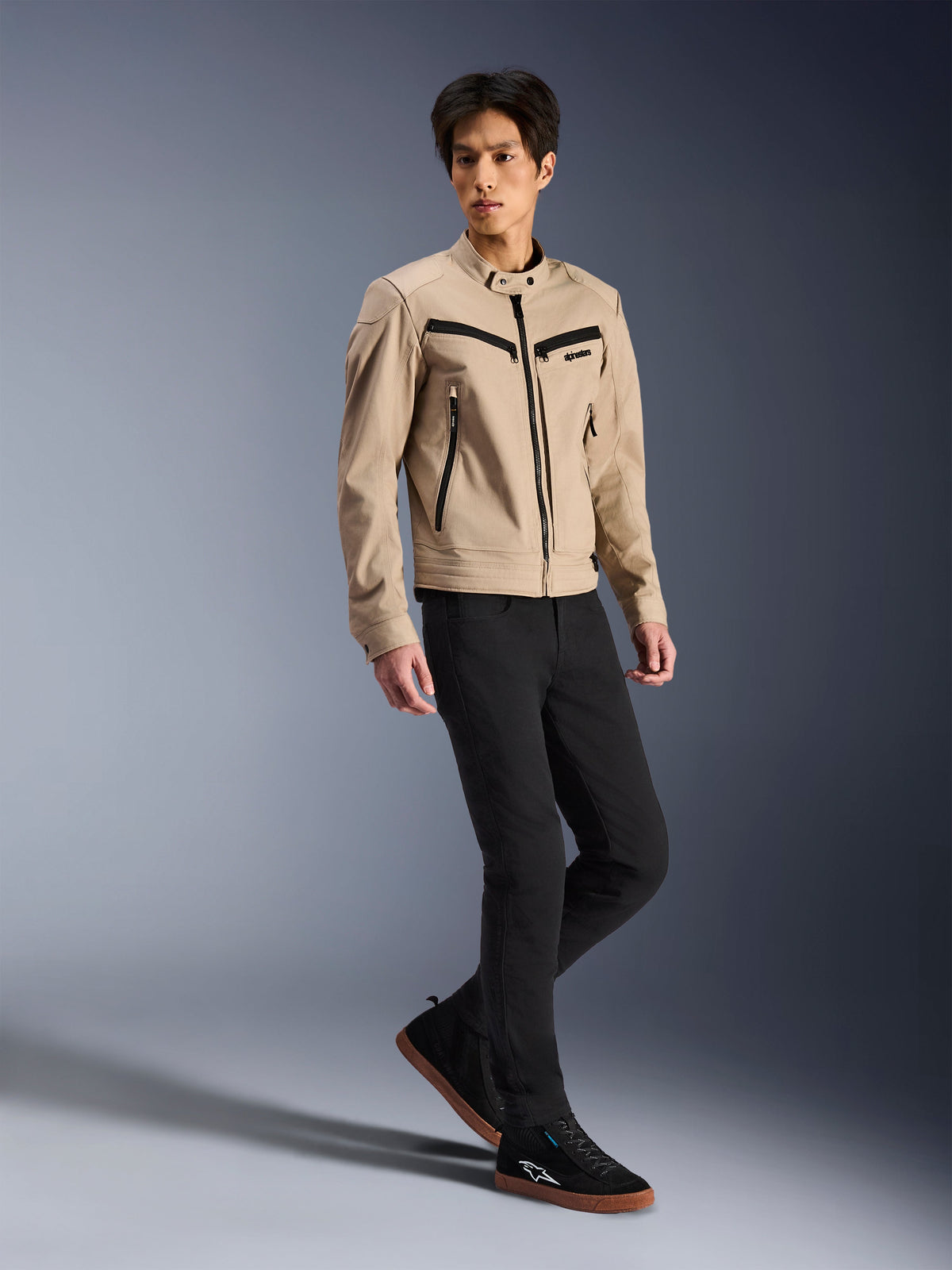 Casaco Alpinestars Bruiser, casaco de motociclismo urbano, bege, vestido por um modelo masculino com calças pretas de corte justo e sapatilhas pretas, construção têxtil inspirada em vestuário de trabalho com detalhes de fechos pretos, estilo urbano contemporâneo.